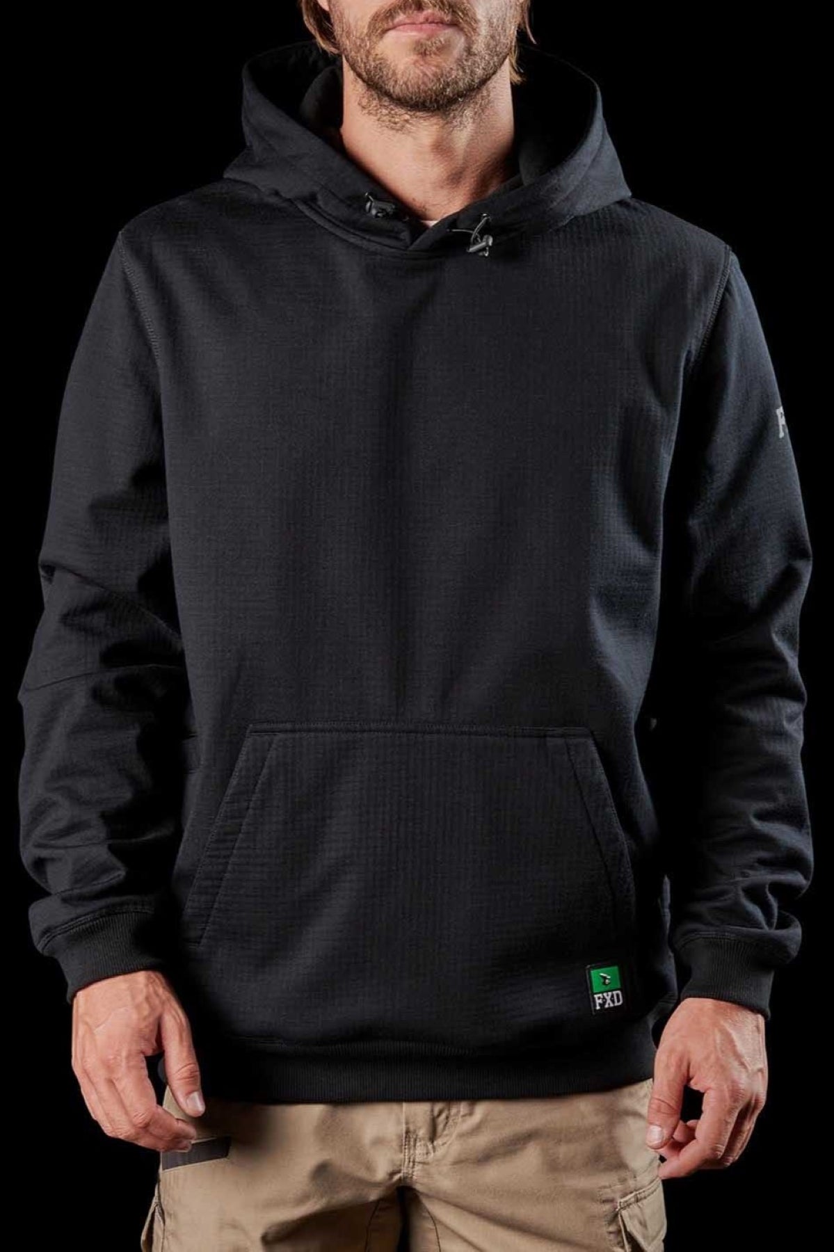 A person is wearing the FXD WF-1 Hoodie, which is black with a front pocket and a logo tag on the bottom. It features DURA500™ canvas elbow patches for added durability. They are also dressed in beige pants. The background is black, emphasizing the clothing, and the person's face is not fully visible.