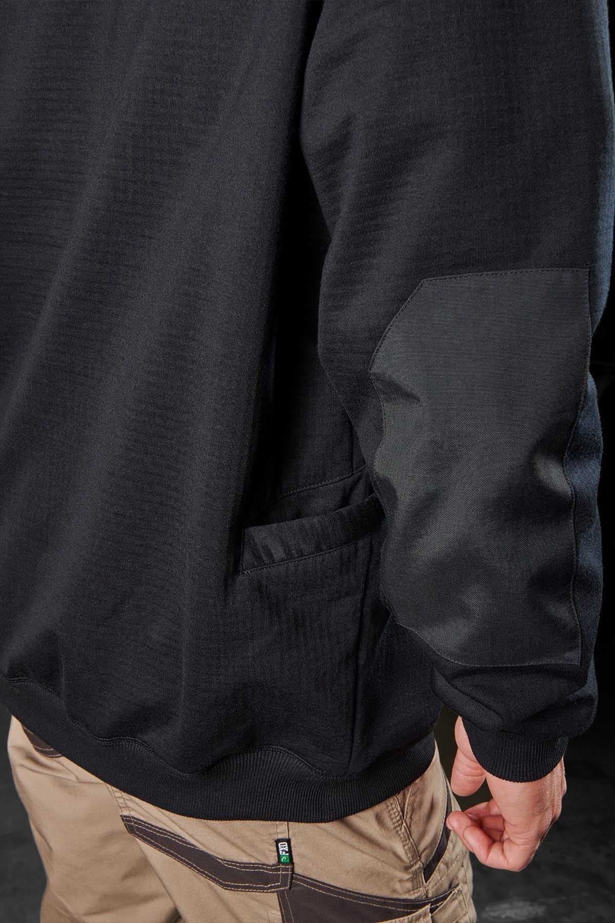 A person is shown from the back, wearing an FXD WF-1 Hoodie adorned with DURA500™ canvas elbow patches and a pair of brown pants featuring a pocket. The hoodie showcases a subtle textured pattern, one visible side pocket, and provides windproof and water-resistant fleece protection. The person is standing against a dark background.