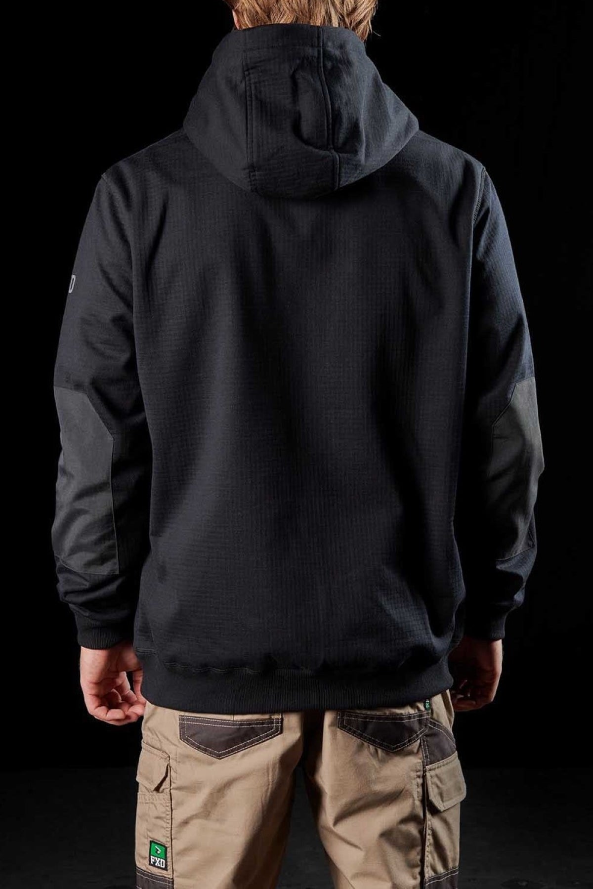 A person with light brown hair stands with their back to the camera, donning an FXD WF-1 Hoodie featuring water-resistant fleece and DURA500™ canvas elbow patches. They are also wearing beige cargo pants adorned with multiple pockets and patches. The solid black background accentuates the clothing's details.