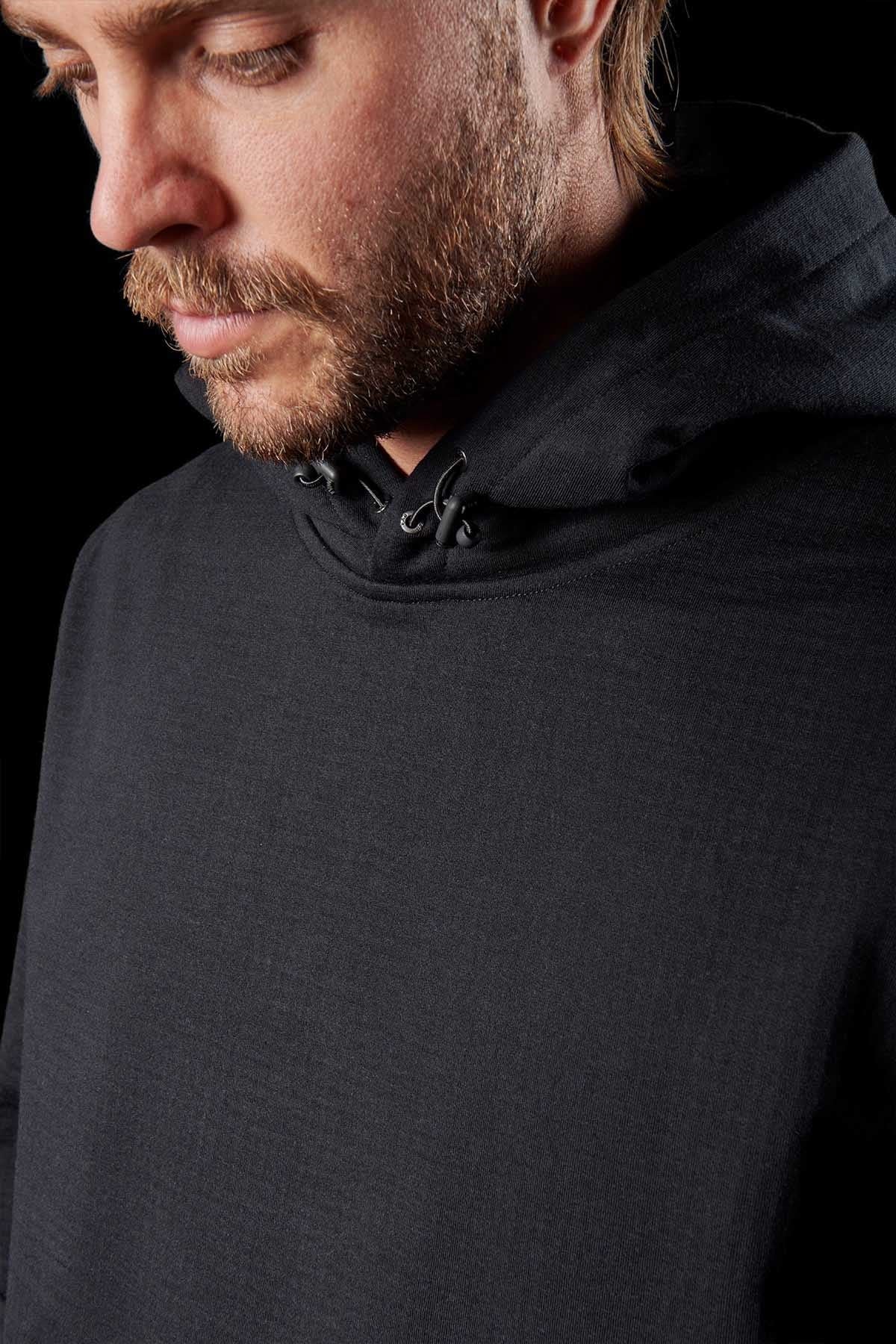 A close-up image portrays a man with short hair and a beard, gazing downward. He is dressed in an FXD WF-1 Hoodie with the hood down, highlighted by DURA500™ canvas elbow patches. The dimly lit background accentuates the man's profile and the hoodie’s fabric texture.