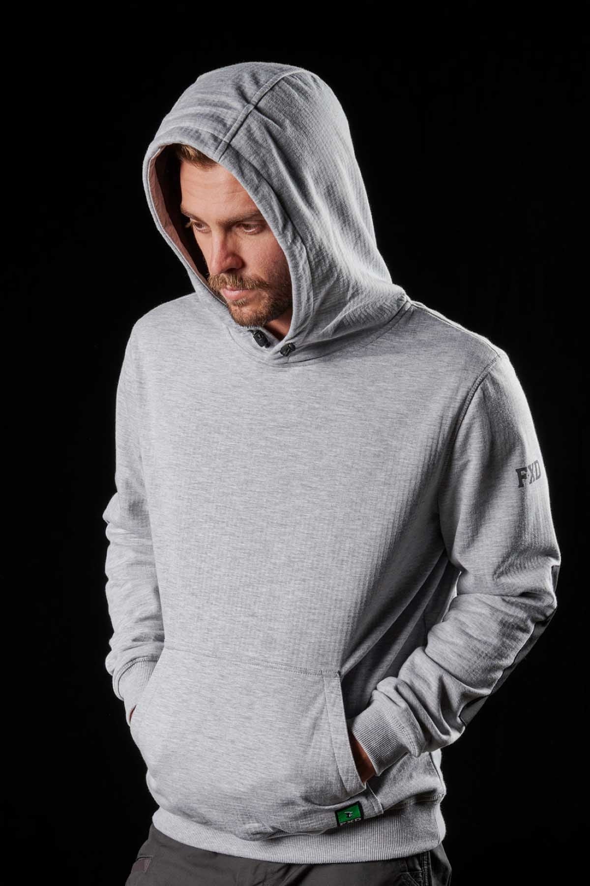 A man with a beard in a grey FXD WF-1 hoodie stands against a black background, looking down with his hands in his pockets. The sleek, water-resistant hoodie features a simple design with a subtle logo on the left sleeve.