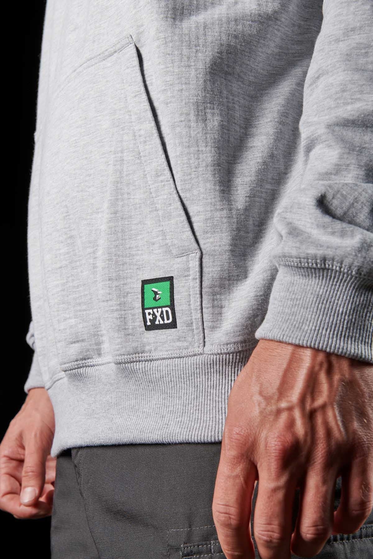 In this close-up, a person is seen wearing the FXD WF-1 Hoodie in light gray, showcasing its windproof fleece material. The hoodie features a small patch near the pocket with the letters "FXD" in black, white, and green. The individual's right hand is visible, and they are dressed in dark pants against a black backdrop.