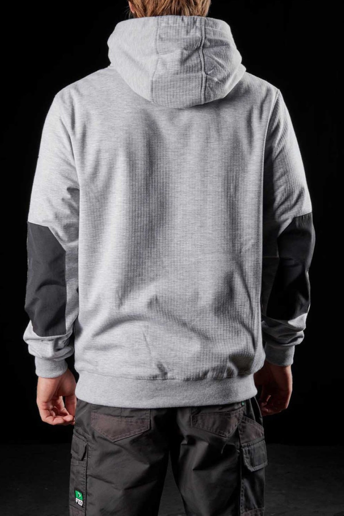 A person stands with their back to the camera, donning an FXD WF-1 Hoodie in light gray, featuring a hood up and black patches on the sleeves. They are also wearing dark-colored pants adorned with a green logo near the pocket. The background is completely black.