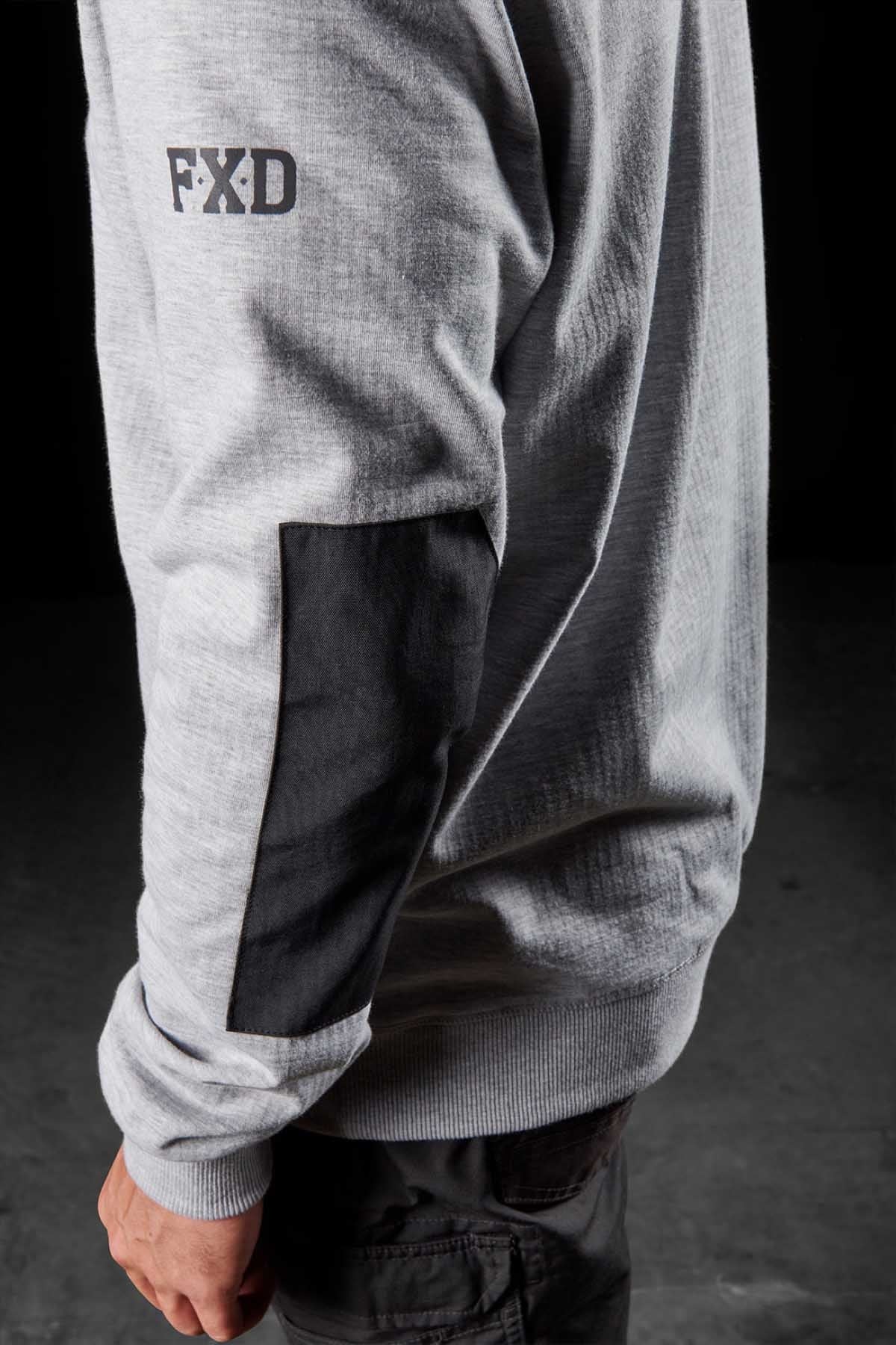 A person wearing the FXD WF-1 Hoodie in light gray, featuring a black patch on the elbow and "FXD" printed on the upper arm, stands against a dark background. Only the right side of their torso and arm is visible. Dressed in dark pants, they look prepared for any weather with their windproof fleece hoodie.