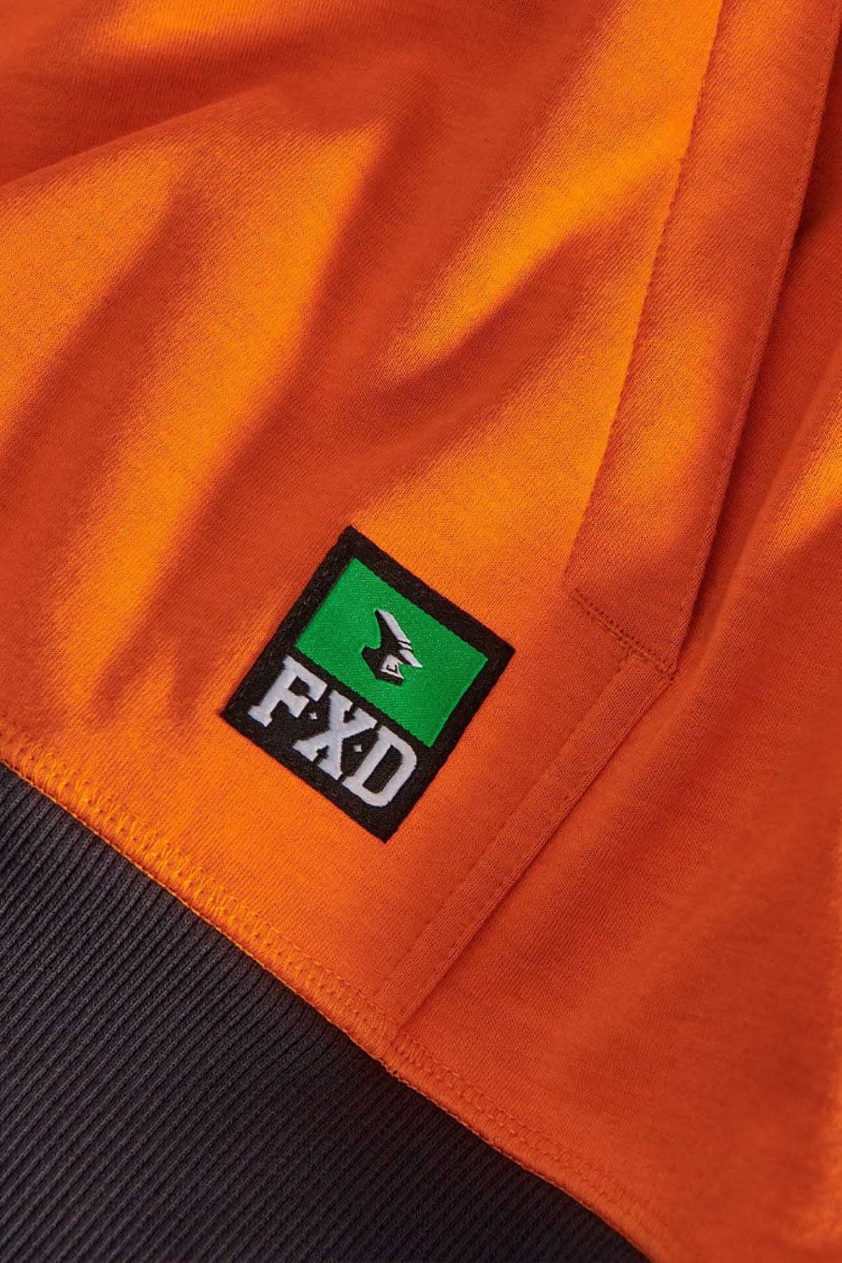 Close-up of an orange fabric from the FXD WF-1 Hoodie, featuring a rectangular patch with a green background, a white bird silhouette, and black text reading "FXD." The material looks durable and practical for various weather conditions.