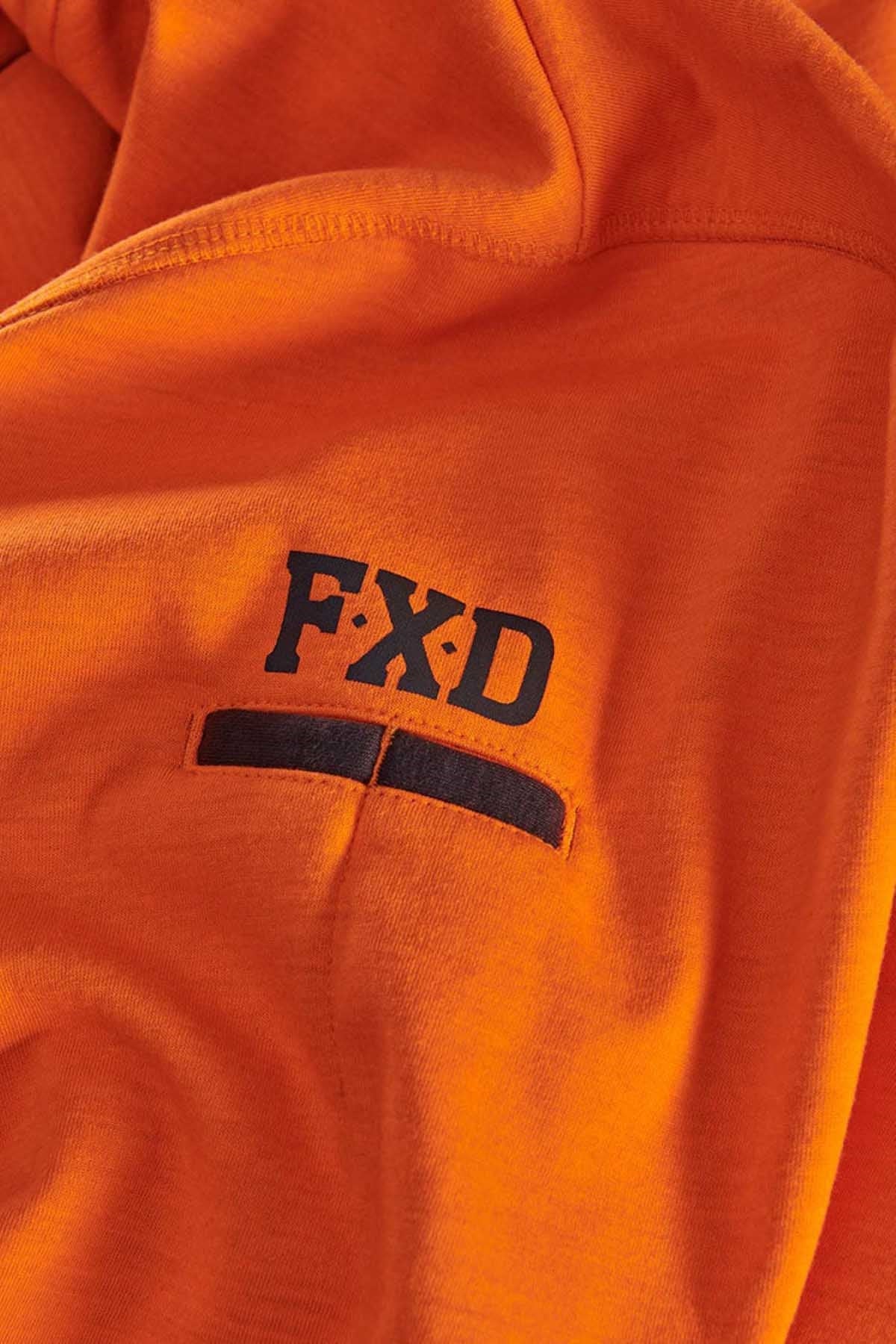 Close-up of an orange fabric with a black "FXD" logo printed on it. The fabric appears to be part of a garment, likely the FXD WF-1 Hoodie, featuring a pocket with a black horizontal band beneath the logo.