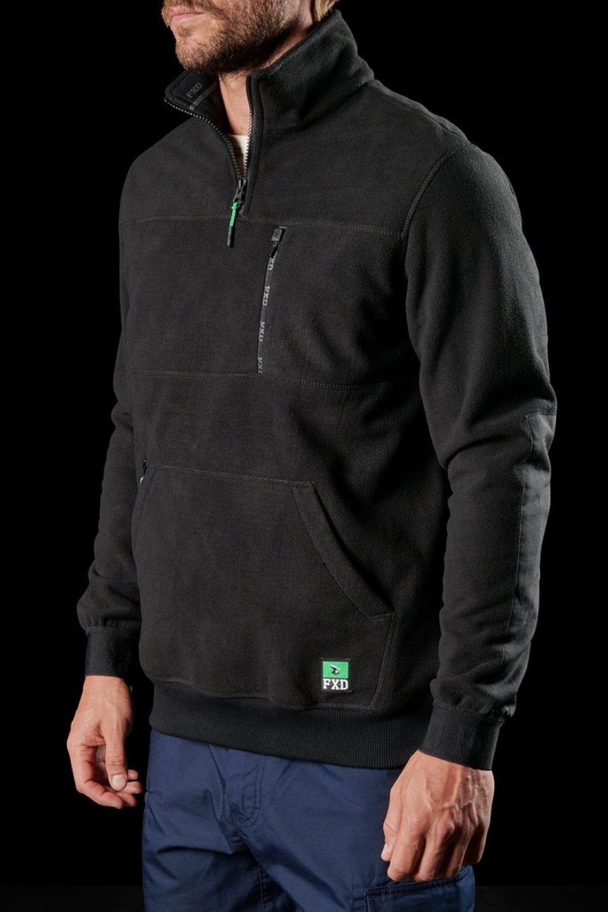A man stands against a black background wearing the FXD WF-2 Fleece—black, windproof, with a quarter zip, zippered chest pocket, front pouch pocket, and a green PXD logo patch near the hem.
