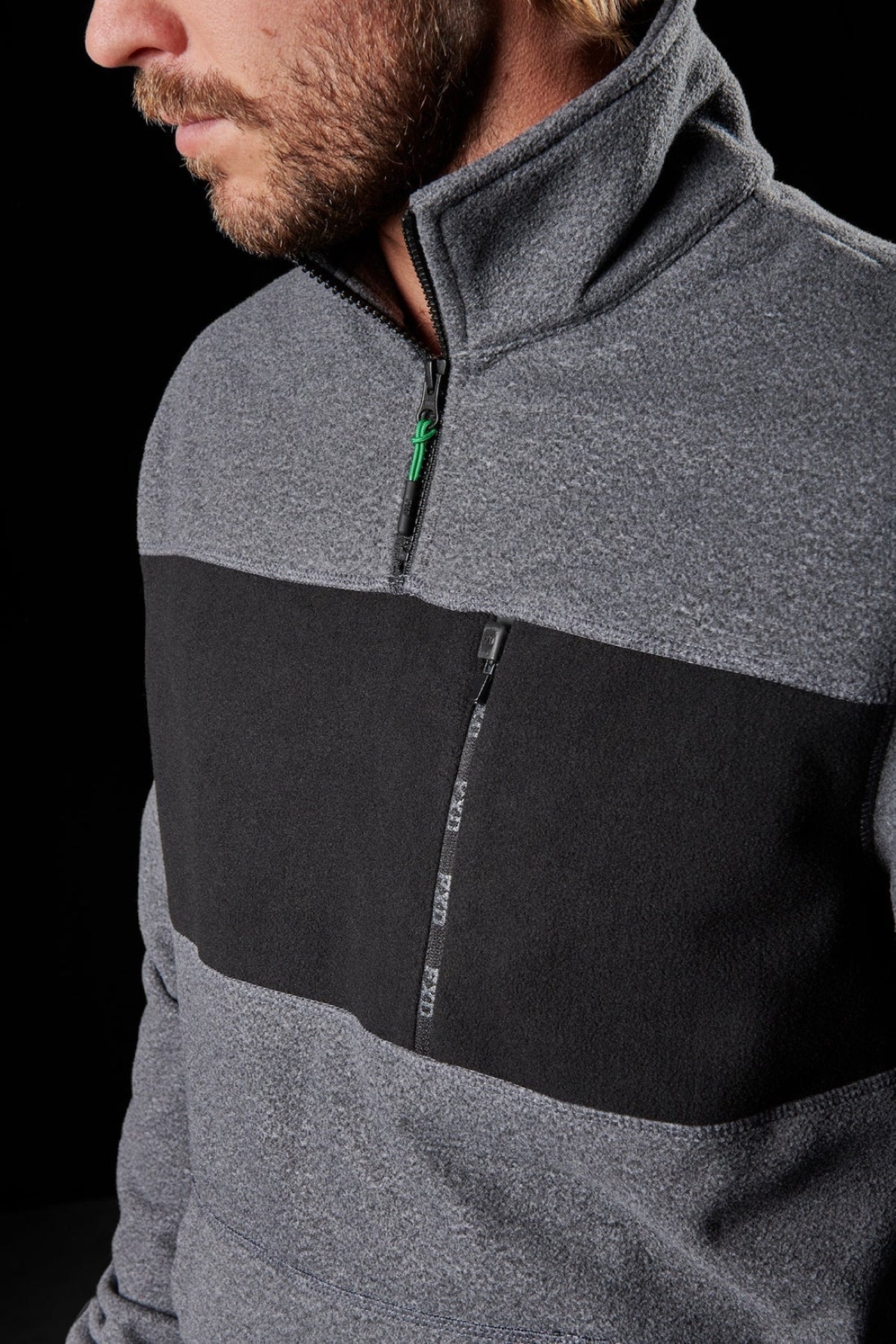 A close-up shows a man wearing the FXD WF-2 Fleece, a gray windproof quarter-zip with a black chest panel and zippered pocket. Only his lower face and upper torso are visible against a black background.
