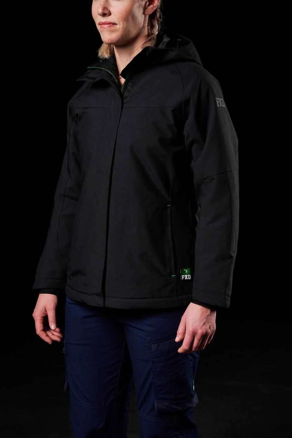A person stands against a black background, wearing the FXD WO-1W Womens Insulated Work Jacket and dark blue pants. They face slightly left, with their face partially visible and hands relaxed at their sides.
