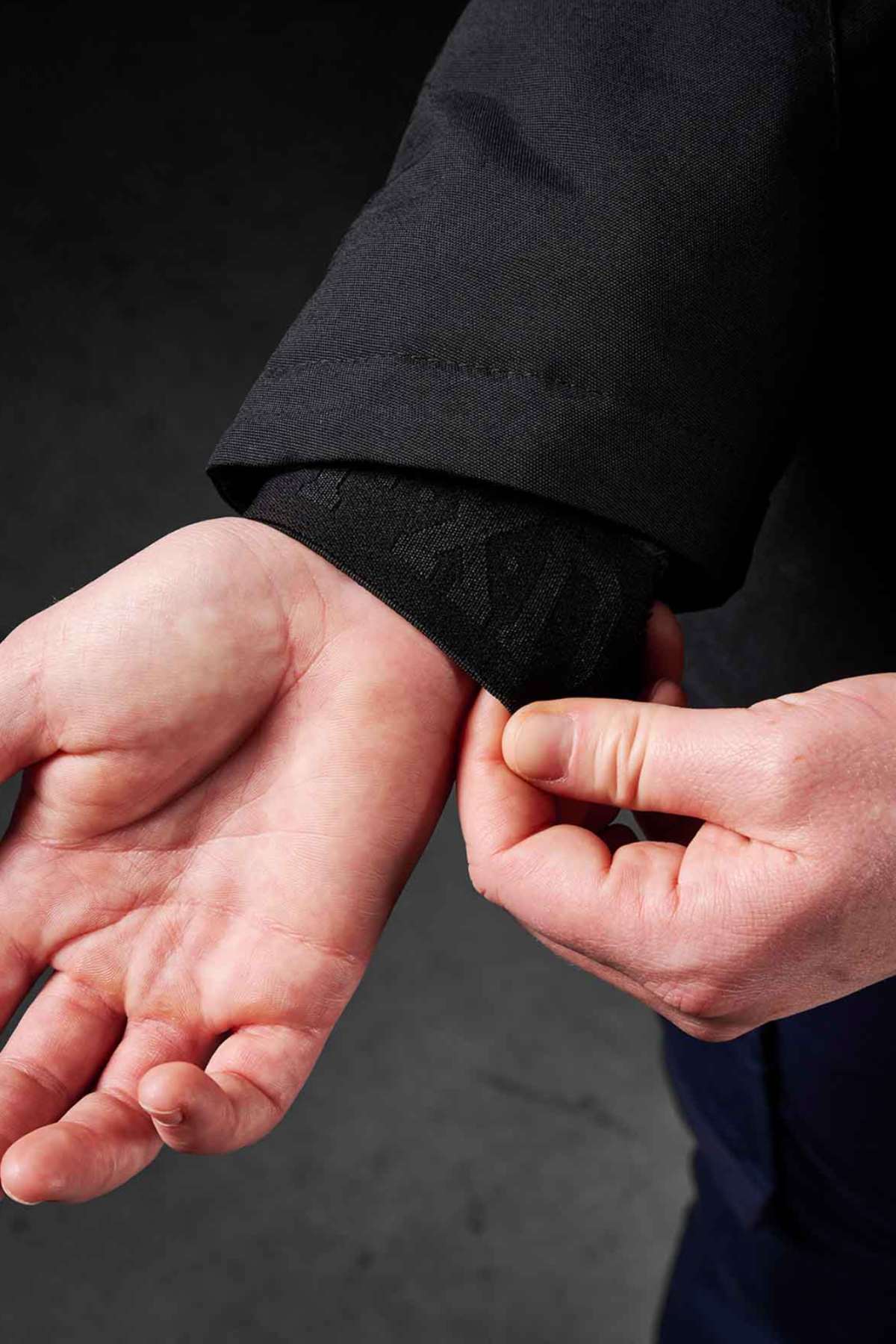 A close-up shows a person adjusting the cuff of the FXD WO-1W Womens Insulated Work Jacket with one hand, while the other hand is open, revealing the wrist and forearm against a dark background.