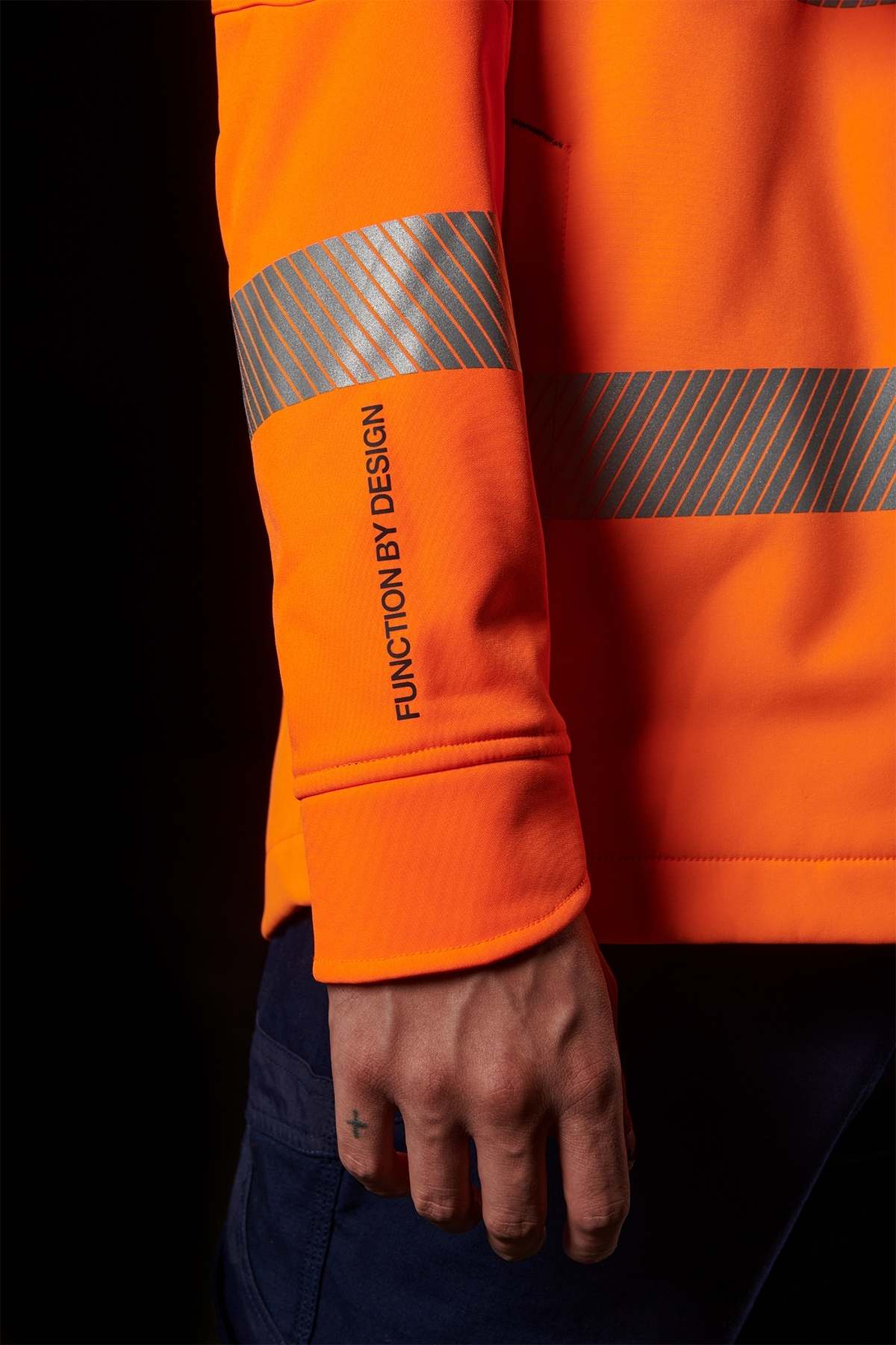 A person wearing an FXD WO-3T Hi Vis Soft Shell Work Jacket with reflective stripes stands against a black background. Featuring a durable water repellent finish, the jacket's sleeve boasts "FUNCTION BY DESIGN" in bold print. The person has a small cross tattoo on their index finger.