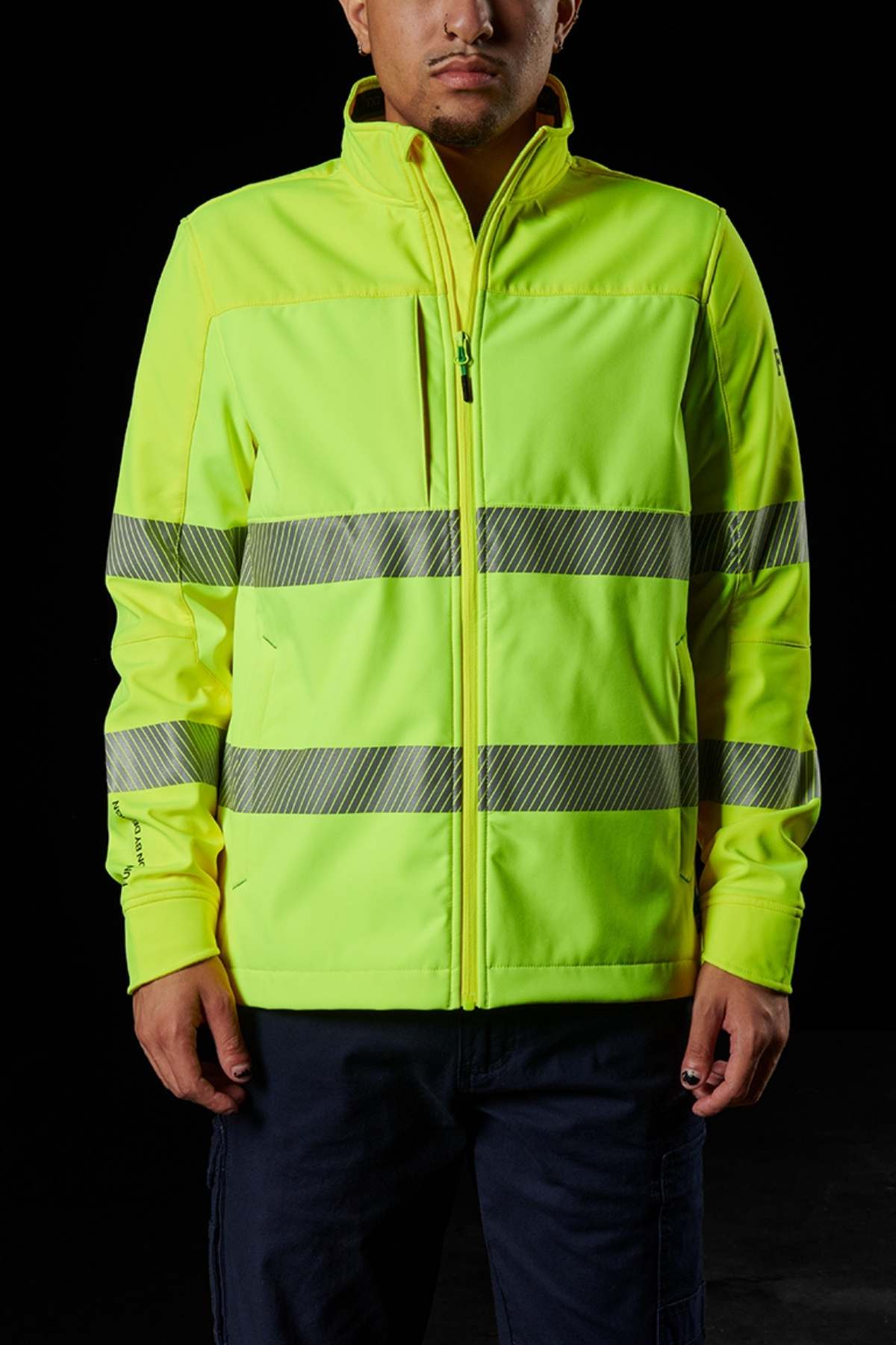 A person is wearing an FXD WO-3T Hi Vis Soft Shell Work Jacket in bright yellow with reflective stripes, offering UPF protection and resisting wind and water, set against a dark background.