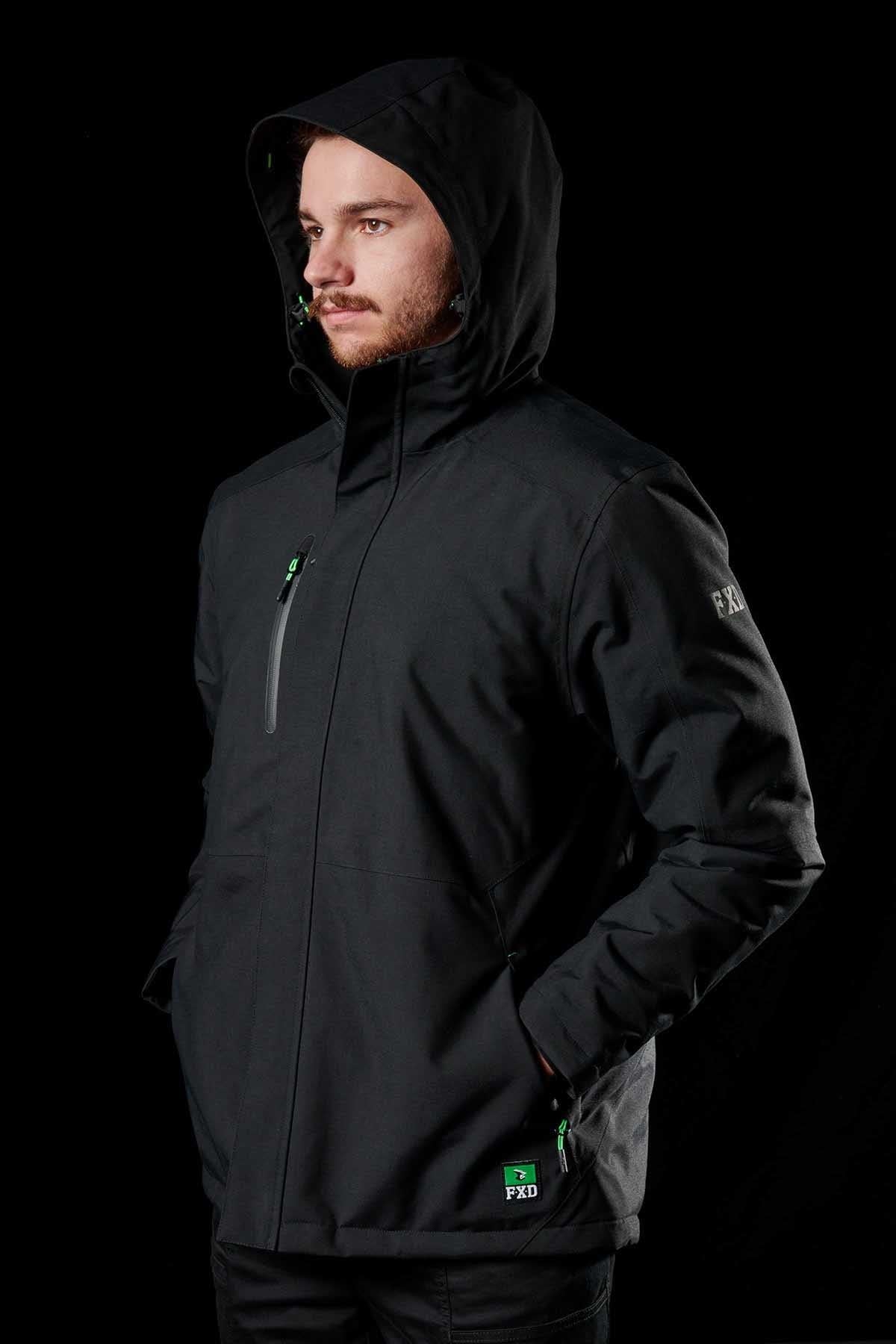 A man stands against a black background wearing the FXD WO-1 Insulated Work Jacket, featuring black fabric, green zipper accents, and the "FXD" logo on the sleeve and pocket. His hands are in the pockets as he looks to the side.