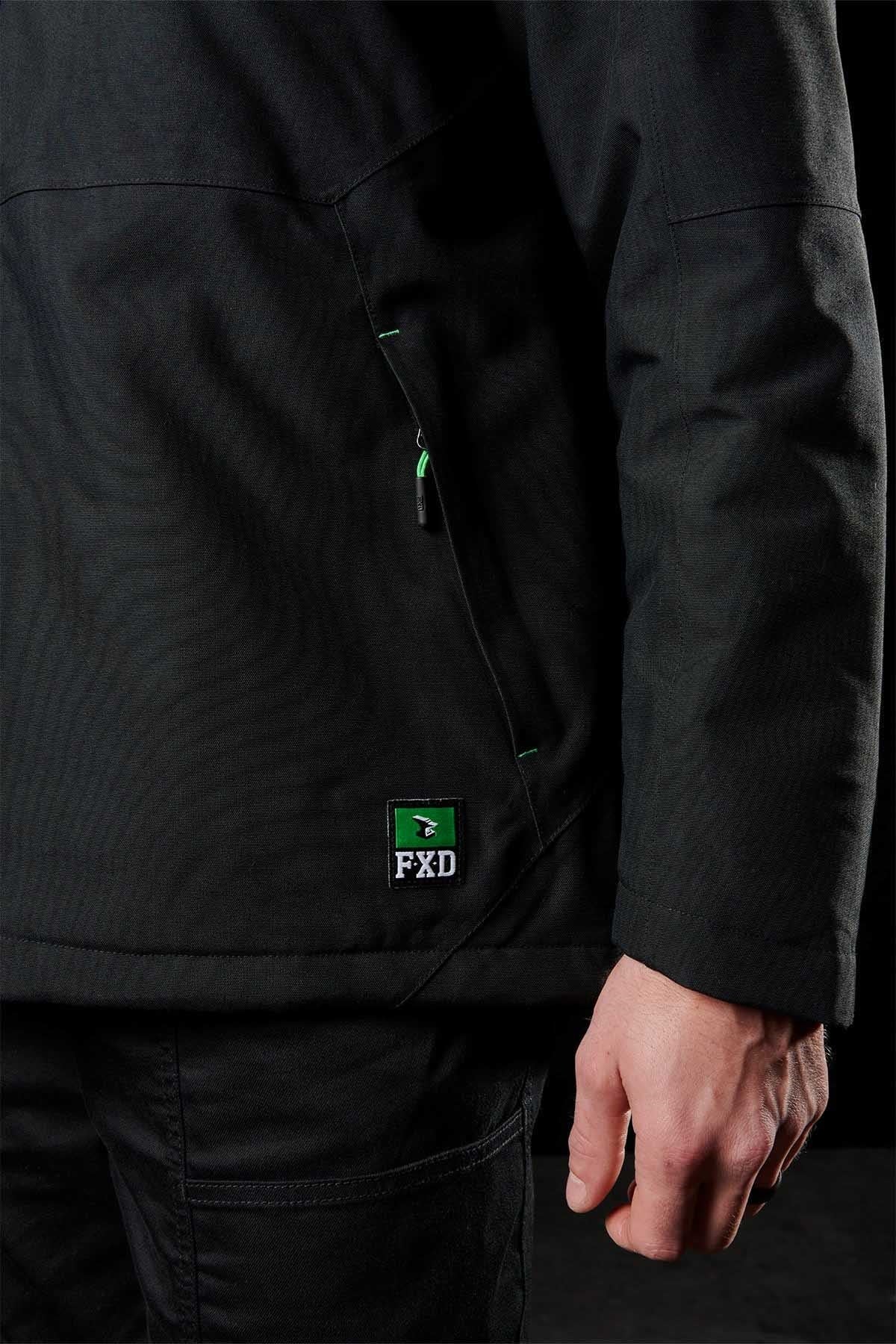 A person wearing a black FXD WO-1 Insulated Work Jacket and black pants stands with their right hand by their side. The FXD logo patch is visible on the lower left of the jacket, near a zippered pocket.