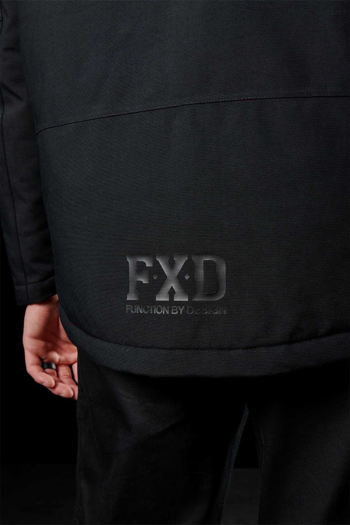 A close-up of the lower back of a person wearing a black FXD WO-1 Insulated Work Jacket with an embossed “FXD FUNCTION BY DESIGN” logo. The person's left hand is visible by their side against a dark background.