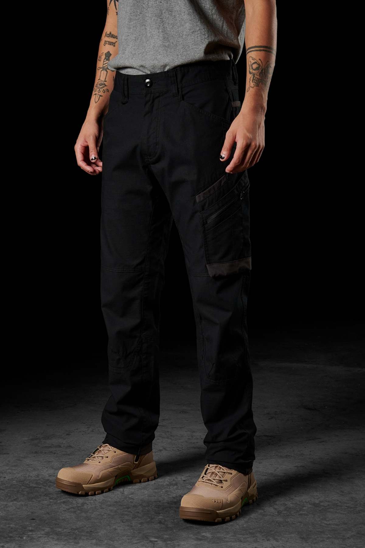 A person wearing black FXD WP-10 work pants with multi-functional cargo pockets, a gray t-shirt, and tan lace-up boots stands against a dark background. Tattoos are visible on their arms.