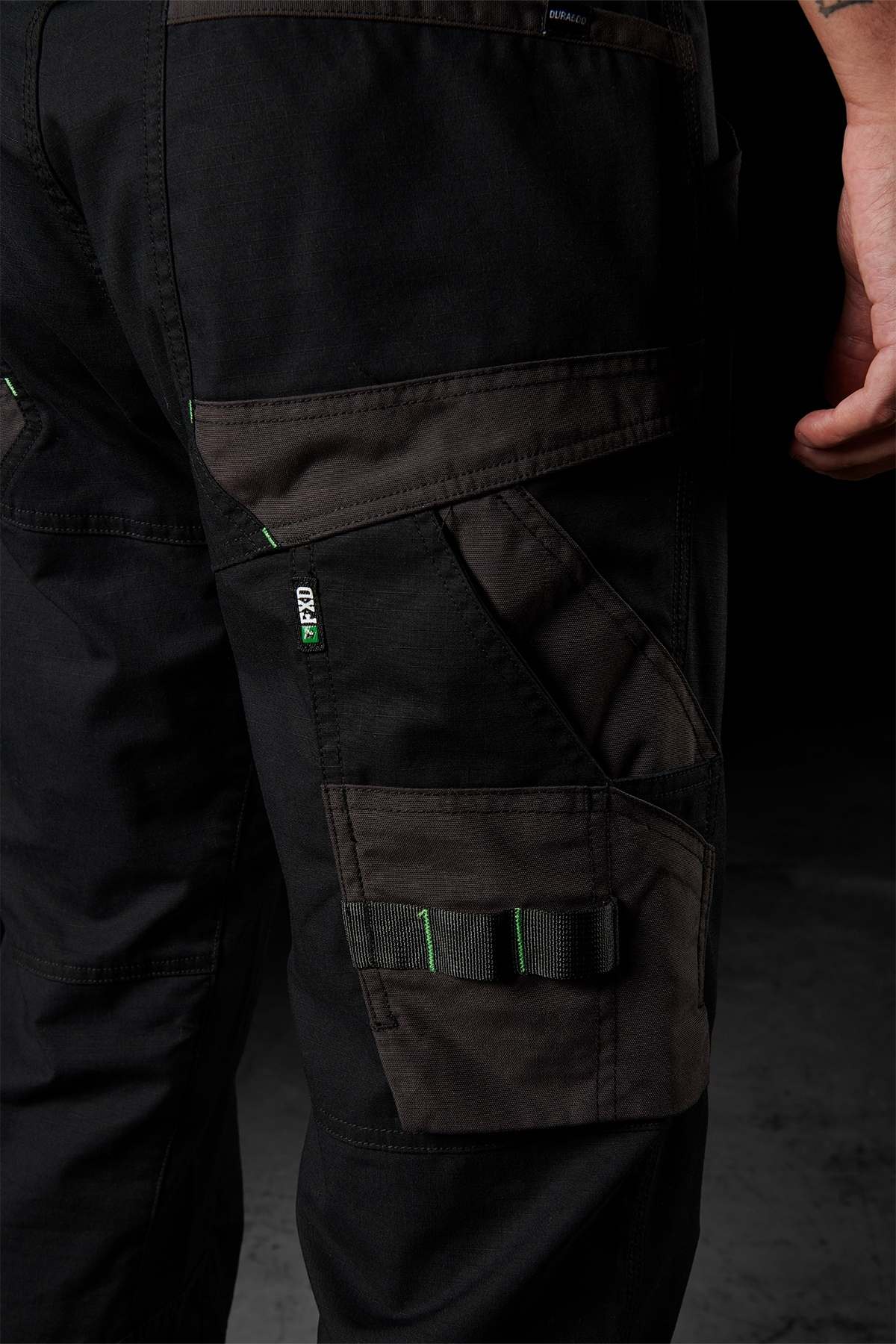 A close-up shot features a person's leg adorned in FXD WP-10 pants, showcasing black, multi-functional cargo pockets with adjustable straps and green threading. The 2-way stretch fabric provides enhanced comfort. The individual stands against a dark background, with their hand partially visible on the right side of the image.