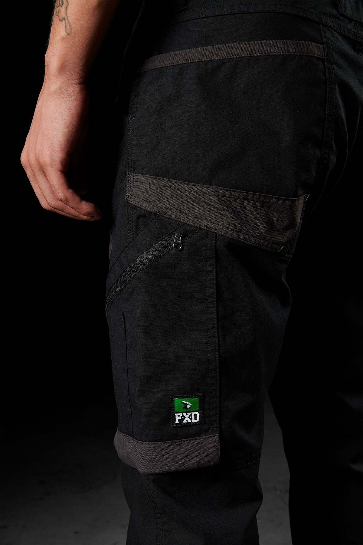 A close-up shows a person wearing FXD WP-10 pants adorned with grey and green accents. The pant leg, crafted from 2-way stretch fabric, prominently displays a green and white "FXD" logo patch above a zipper pocket on the thigh. The background is dark, and the person's partially tattooed left arm is visible.