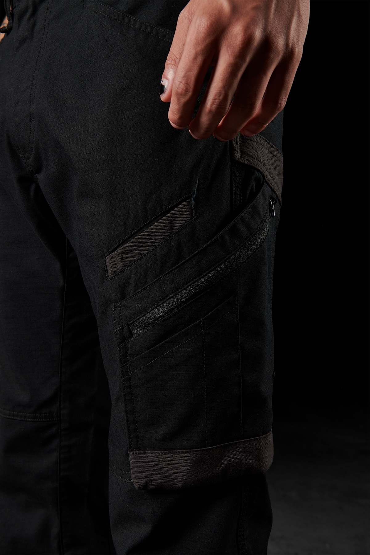 Close-up of a person wearing FXD WP-10 pants with multi-functional cargo pockets, including a zippered pocket on the side. The person's partially visible hand rests near the top pocket. The dark background accentuates the pants' details and 2-way stretch fabric.