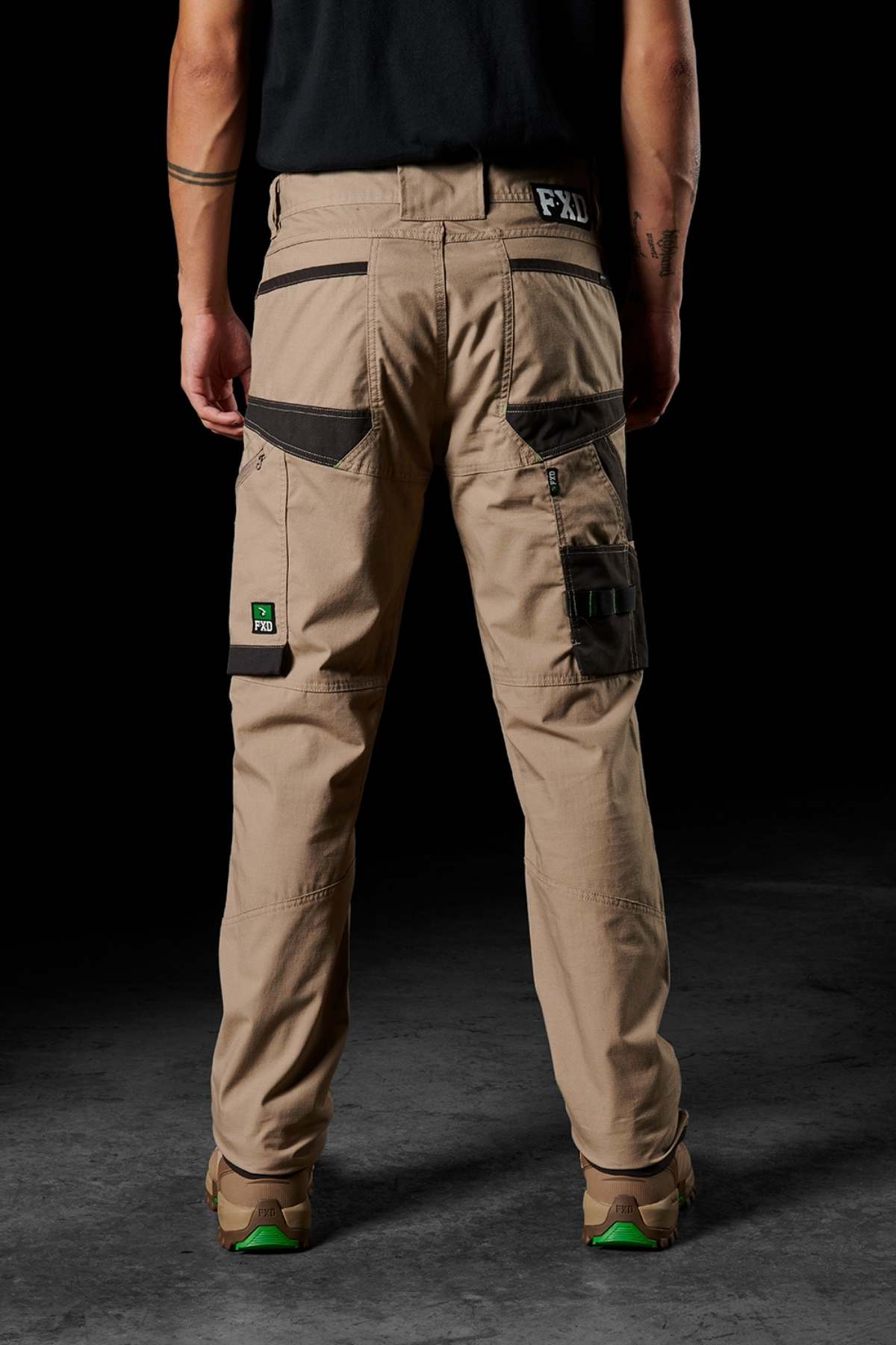 A person stands with their back to the camera, dressed in FXD WP-10 beige pants that include multi-functional cargo pockets and a tool loop. The distinctive brand logo is visible on the right back pocket of the pants. They also wear tan and green work boots along with a black shirt.