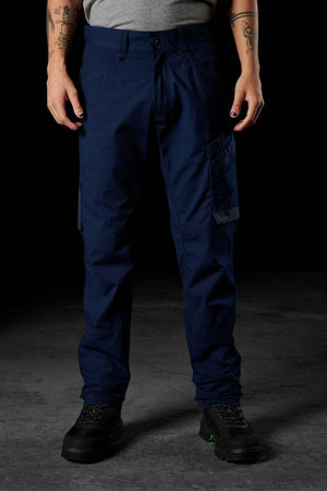 A person stands against a dark background, wearing navy blue FXD WP-10 pants made from 2-way stretch fabric, along with a gray t-shirt and black work boots featuring green accents. Their tattooed hands with painted nails rest at their sides, while the upper body is not visible in the image.