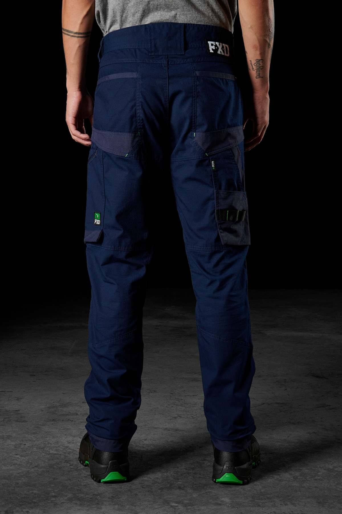 A person wearing dark blue FXD WP-10 pants and black-green shoes is standing in a dimly lit environment. The FXD WP-10 pants feature multi-functional cargo pockets and utility loops, crafted from 2-way stretch fabric. The person's back is facing the camera, showcasing the detailed design of the pants.