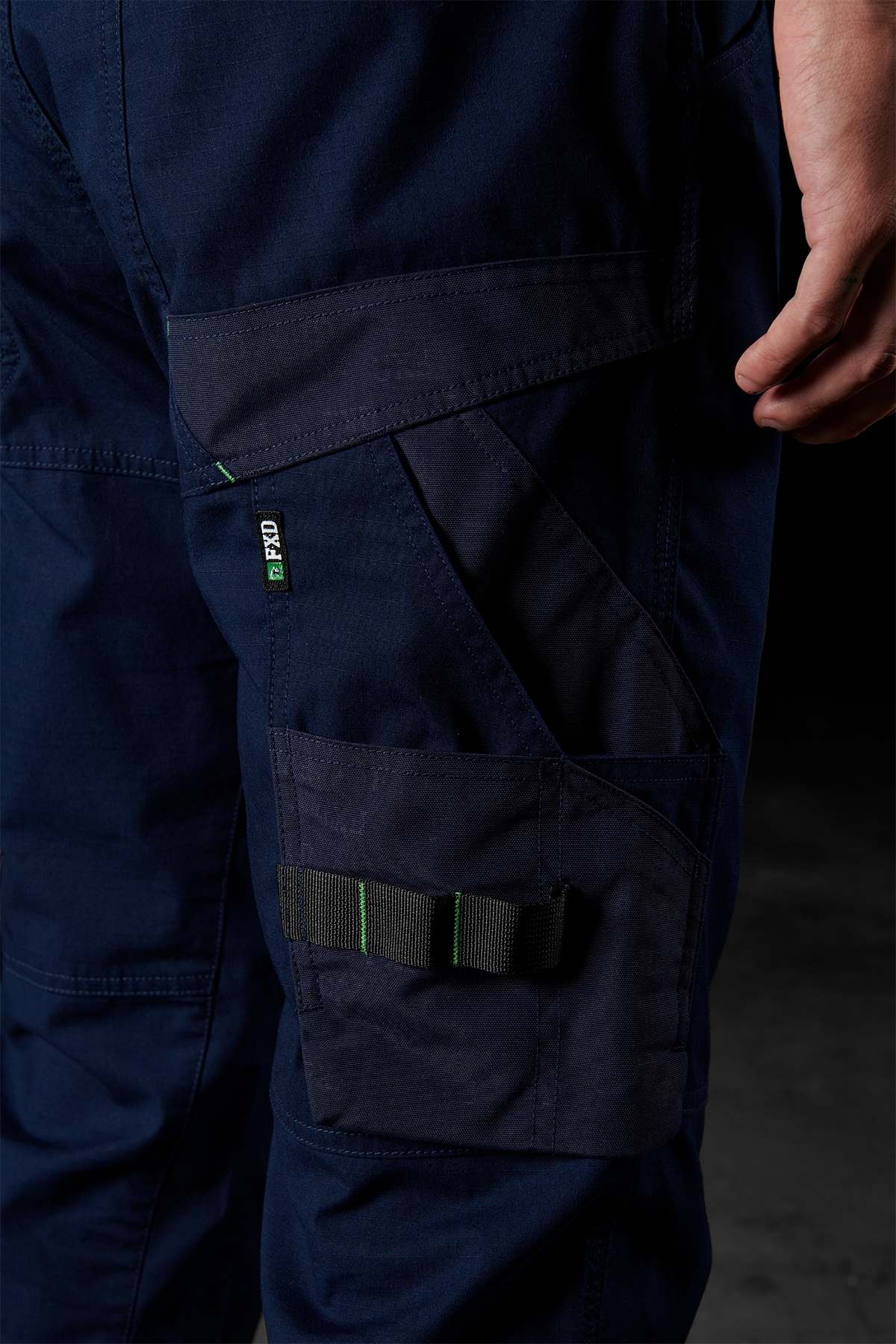 Close-up of a person wearing FXD WP-10 in dark blue, highlighting a side cargo pocket with a green-accented black strap and stitched detailing. The multi-functional cargo pockets are enhanced by the 2-way stretch fabric. The individual stands against a dark background with their right hand partially visible.