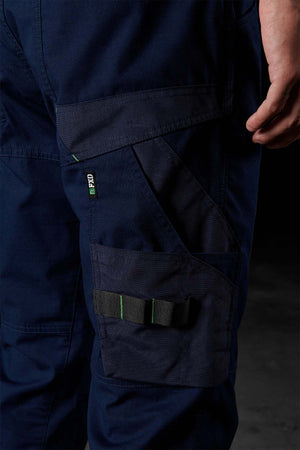 Close-up of a person wearing FXD WP-10 in dark blue, highlighting a side cargo pocket with a green-accented black strap and stitched detailing. The multi-functional cargo pockets are enhanced by the 2-way stretch fabric. The individual stands against a dark background with their right hand partially visible.