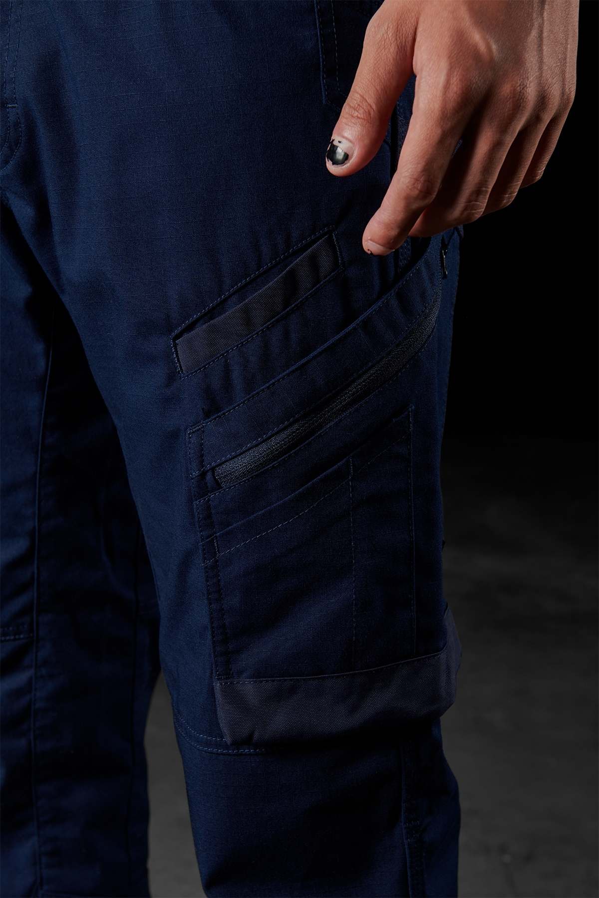 Close-up of a person wearing dark blue FXD WP-10 work pants, featuring multi-functional cargo pockets. The person's hand, adorned with black nail polish, is partially visible resting next to one of the pockets. The background is dark and blurred.