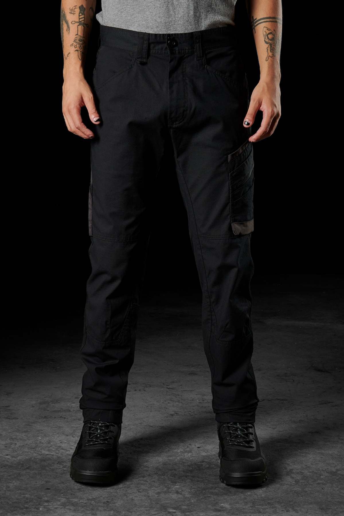 A person is standing against a dark backdrop, dressed in black FXD WP-11 pants featuring multi-functional cargo pockets, black boots, and a grey t-shirt. Tattooed arms are on display, with nails painted black. Their face remains out of view."""

