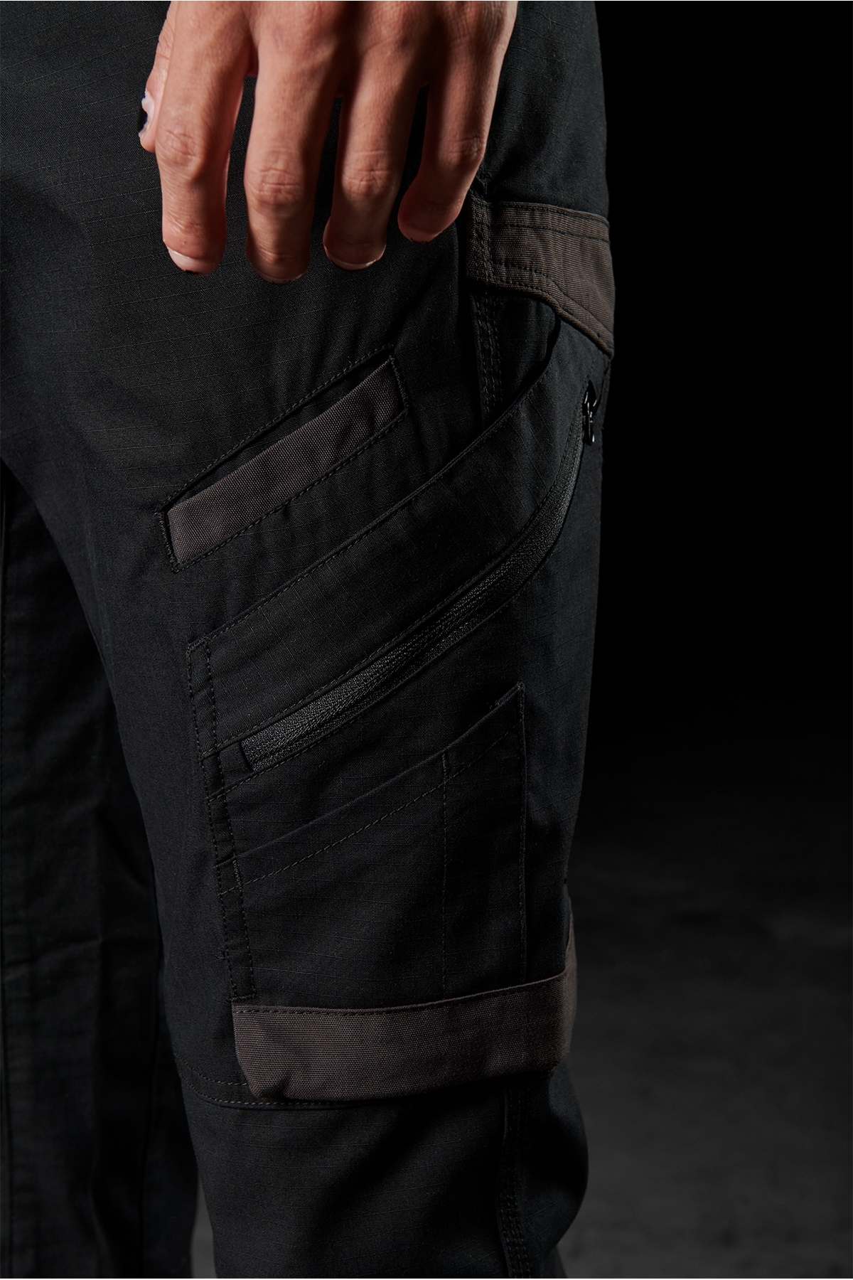 A close-up image shows a person's hand resting on their thigh, clad in FXD WP-11 pants. These black pants, crafted from 2-way stretch fabric, are highlighted by their multi-functional cargo pockets and multiple zippered compartments. The dark and blurred background accentuates the details of both the pants and the hand.