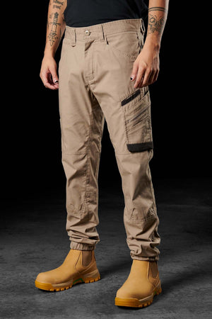 A person wearing beige FXD WP-11 work pants with multi-functional cargo pockets and yellow work boots stands against a black background. Only the lower half is visible, showcasing the detailed stitching and design of the 2-way stretch fabric in the FXD WP-11 pants and the sturdy construction of the boots.