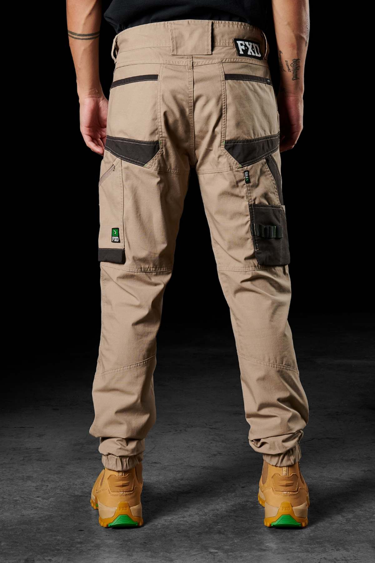 Someone is standing on a dark, textured surface wearing tan FXD WP-11 cargo pants with black accents and multiple pockets. These comfortable pants, labeled "FXD," are made from 2-way stretch fabric. They are also sporting light brown work boots with green rubber soles. Only the lower half of the body is visible.