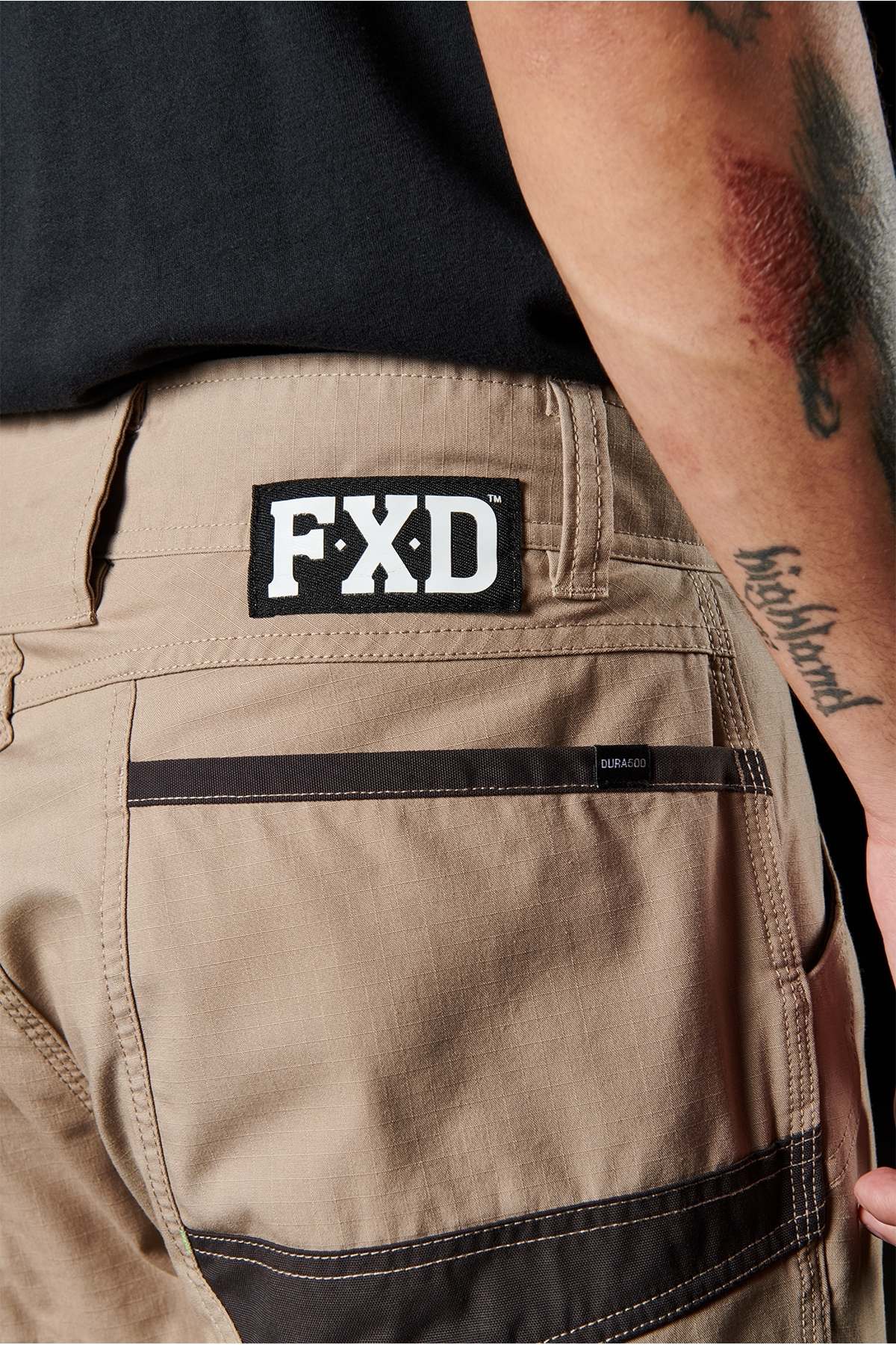 Close-up of a person's hip area wearing FXD WP-11 pants in beige with black detailing and a visible "FXD" tag above the back pocket. The person has a partially visible forearm tattoo. Part of a black shirt is also noticeable, emphasizing the multi-functional cargo pockets and 2-way stretch fabric of the pants.