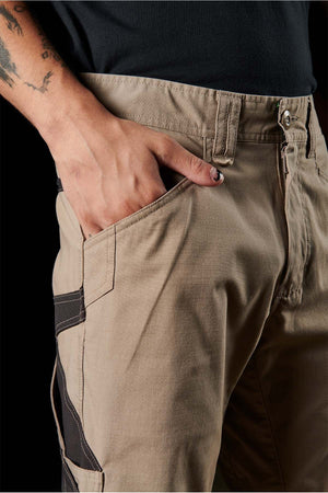 Close-up of a person wearing FXD WP-11 pants with a hand placed in the front pocket. The beige FXD WP-11 pants feature black detailing on the side, multi-functional cargo pockets, and a zipper partially visible at the fly. The person's arm reveals partial tattoos and nail polish on the thumb.