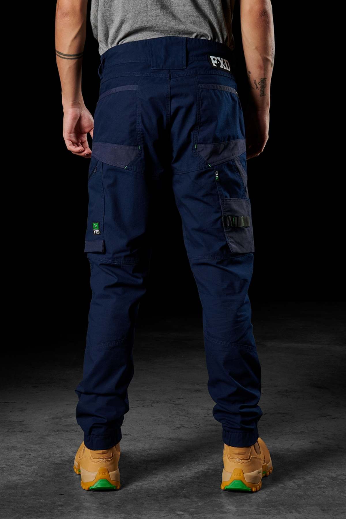 A person wearing dark blue FXD WP-11 work pants with multiple cargo pockets is viewed from the back. The pants feature a small logo on the rear waistband and have lighter blue accents. The individual is also wearing tan work boots and has tattoos on their arms.