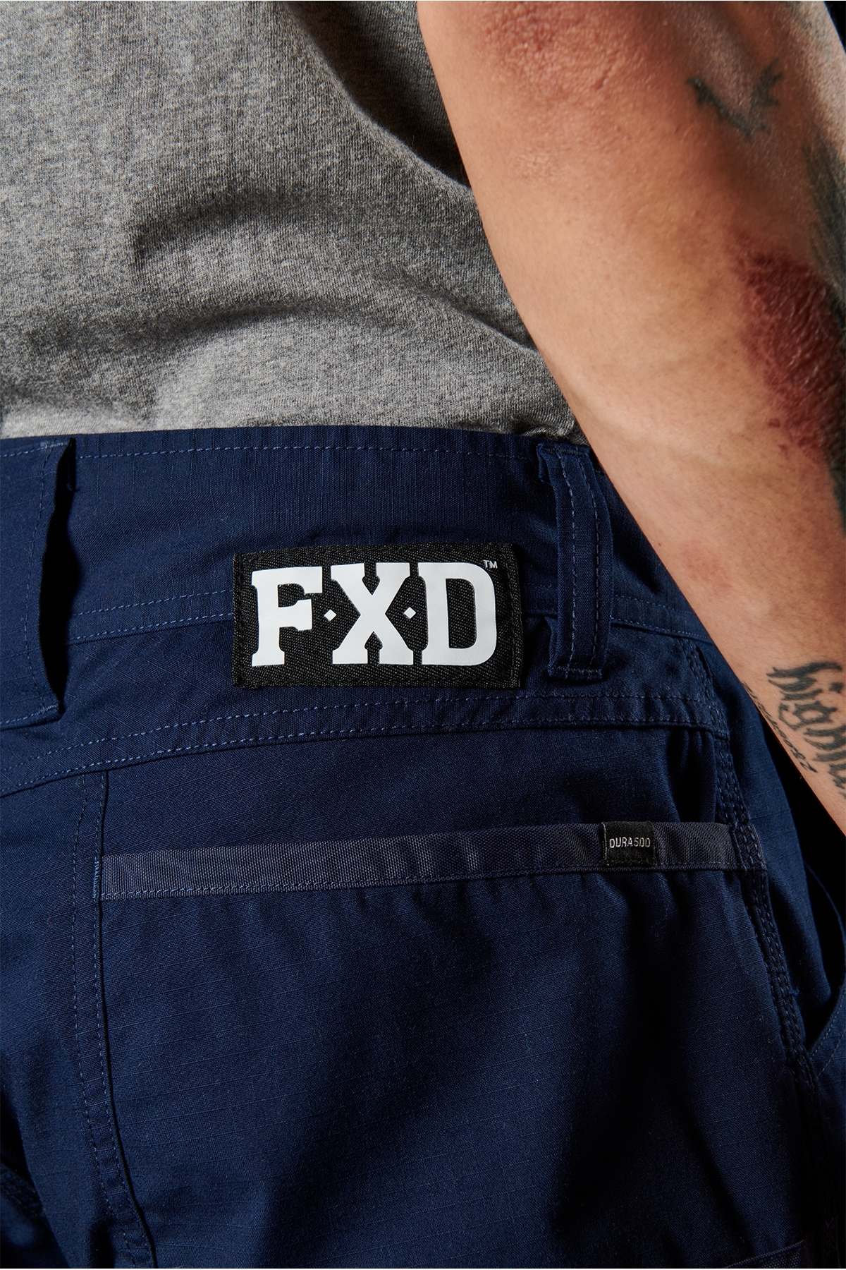 Close-up of a person wearing navy blue FXD WP-11 Work Pants with "FXD" branding on the back pocket. The pants feature cargo pockets and 2-way stretch fabric. The person is also wearing a gray shirt, and their tattooed arm is visible in the frame.