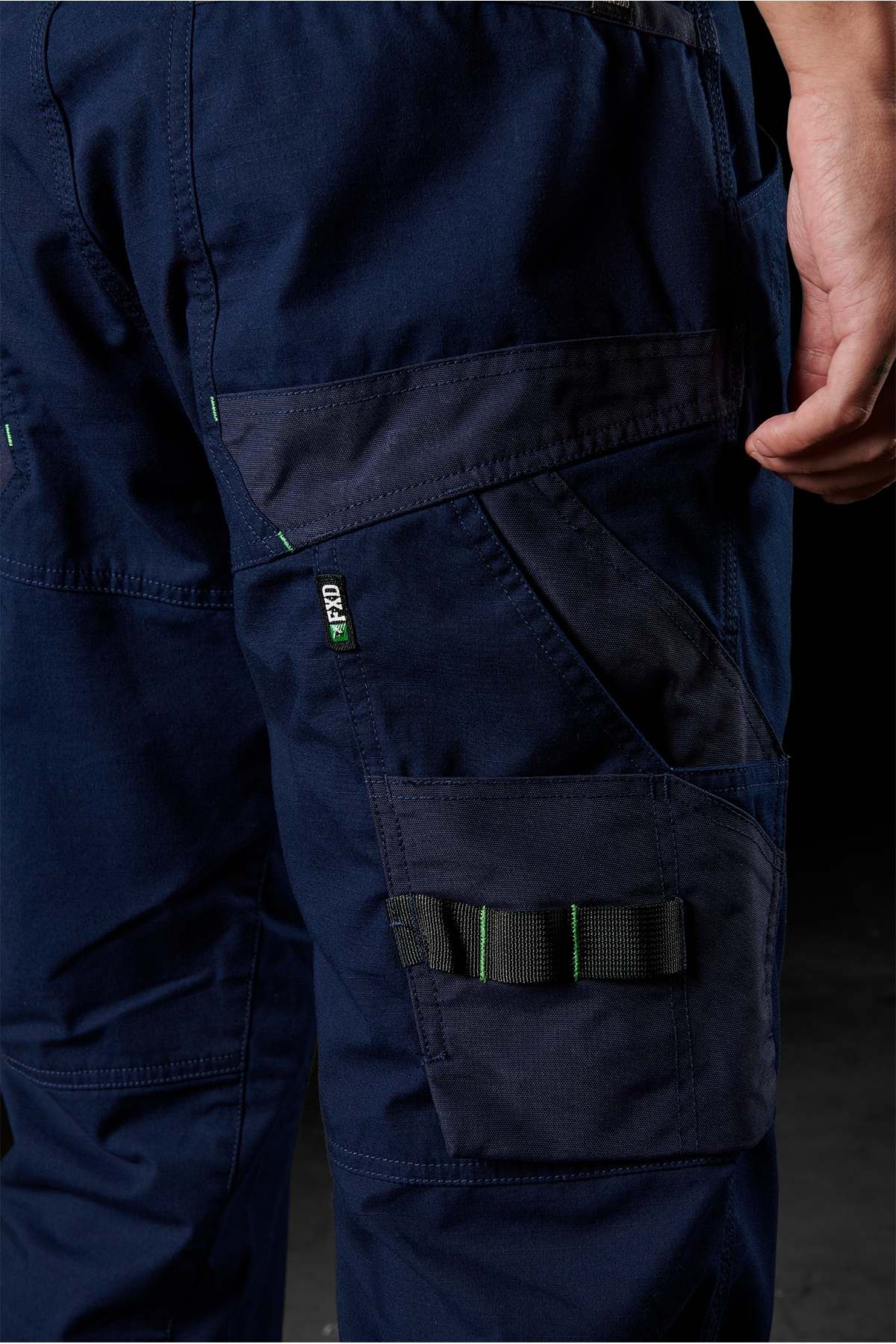 Close-up of a person wearing the FXD WP-11 pants, showcasing their navy blue color, multiple cargo pockets, and distinctive green stitching details. These pants offer various compartments for tools and feature 2-way stretch fabric, all set against a dark background that emphasizes their design and functionality. The person's hand is partially visible.