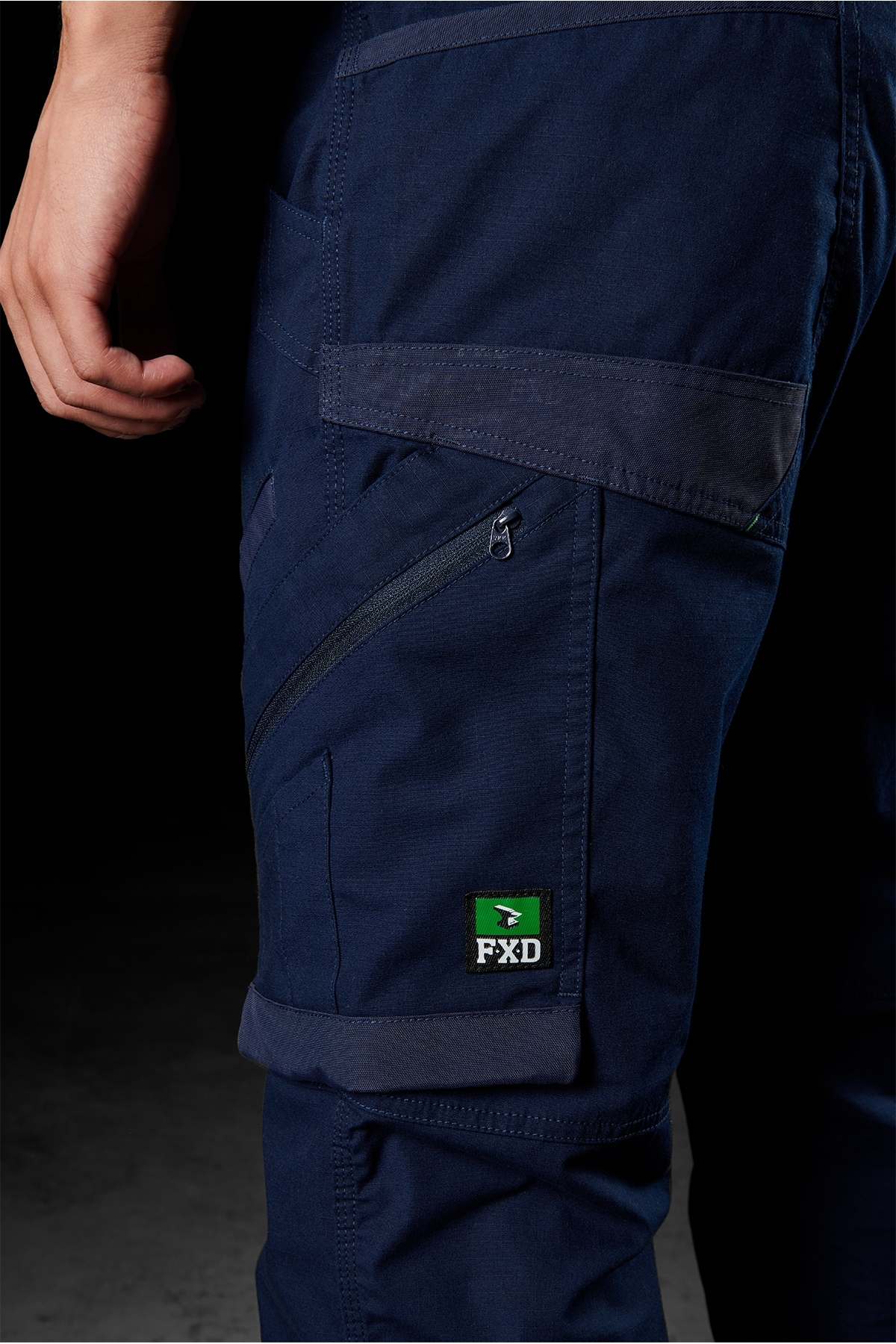 Close-up of a person wearing navy blue FXD WP-11 pants. The cargo pockets feature multiple zippers and display a green FXD logo patch on one of the pockets. Only the lower torso and part of the left arm are visible against a dark background, which accentuates the 2-way stretch fabric.