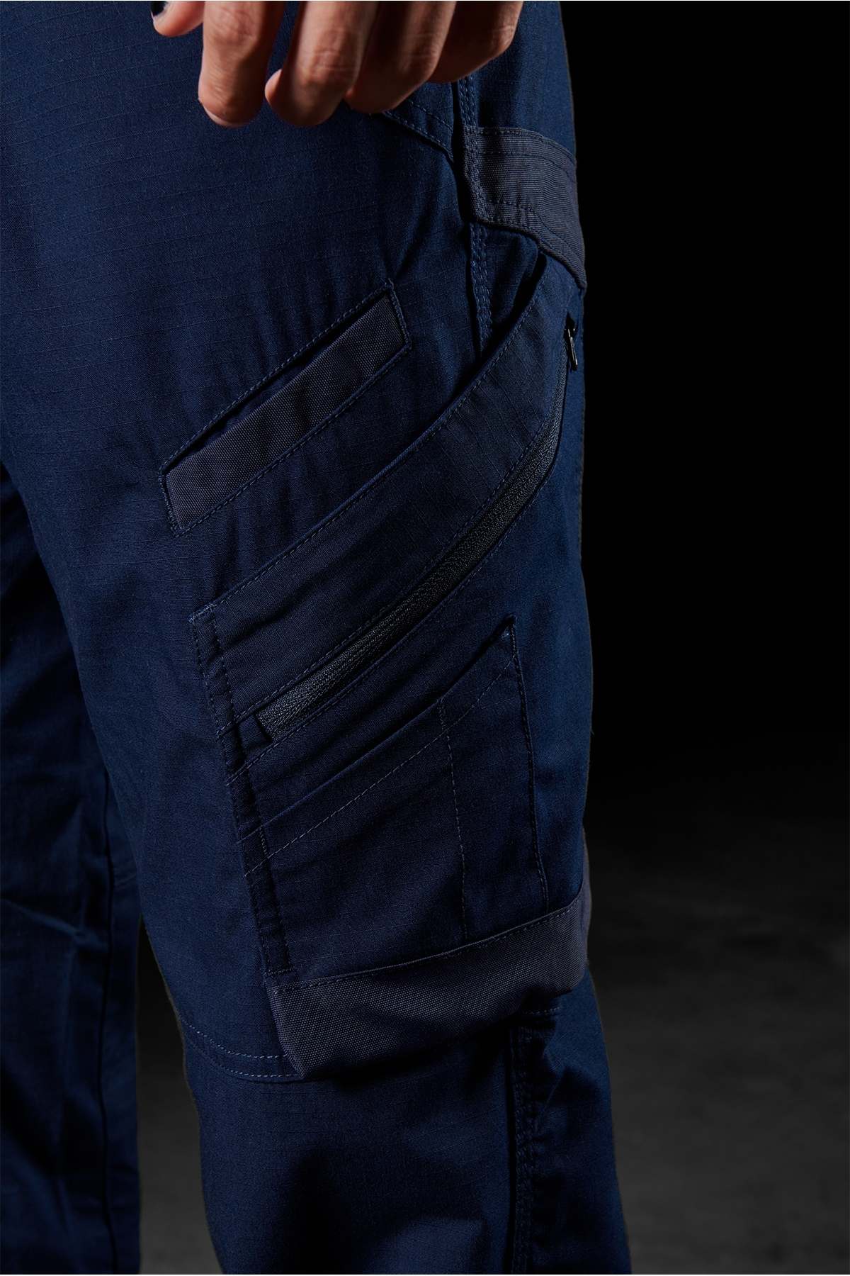 Close-up of a person's hand and a section of their FXD WP-11 pants in blue, highlighting multiple cargo pockets, including one with a zipper on the thigh. The dark background accentuates the details of the 2-way stretch fabric.