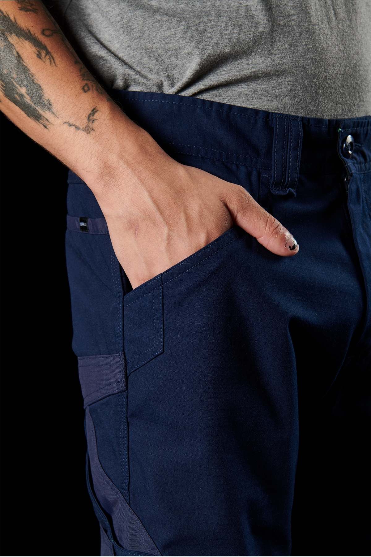 Close-up of a person's hand in the pocket of navy blue FXD WP-11 pants, showcasing the 2-way stretch fabric. The person is wearing a grey shirt and displaying a tattoo on their forearm. The cargo pocket features a flap and intricate stitching detail, and the person's short fingernails are painted. The background is black.