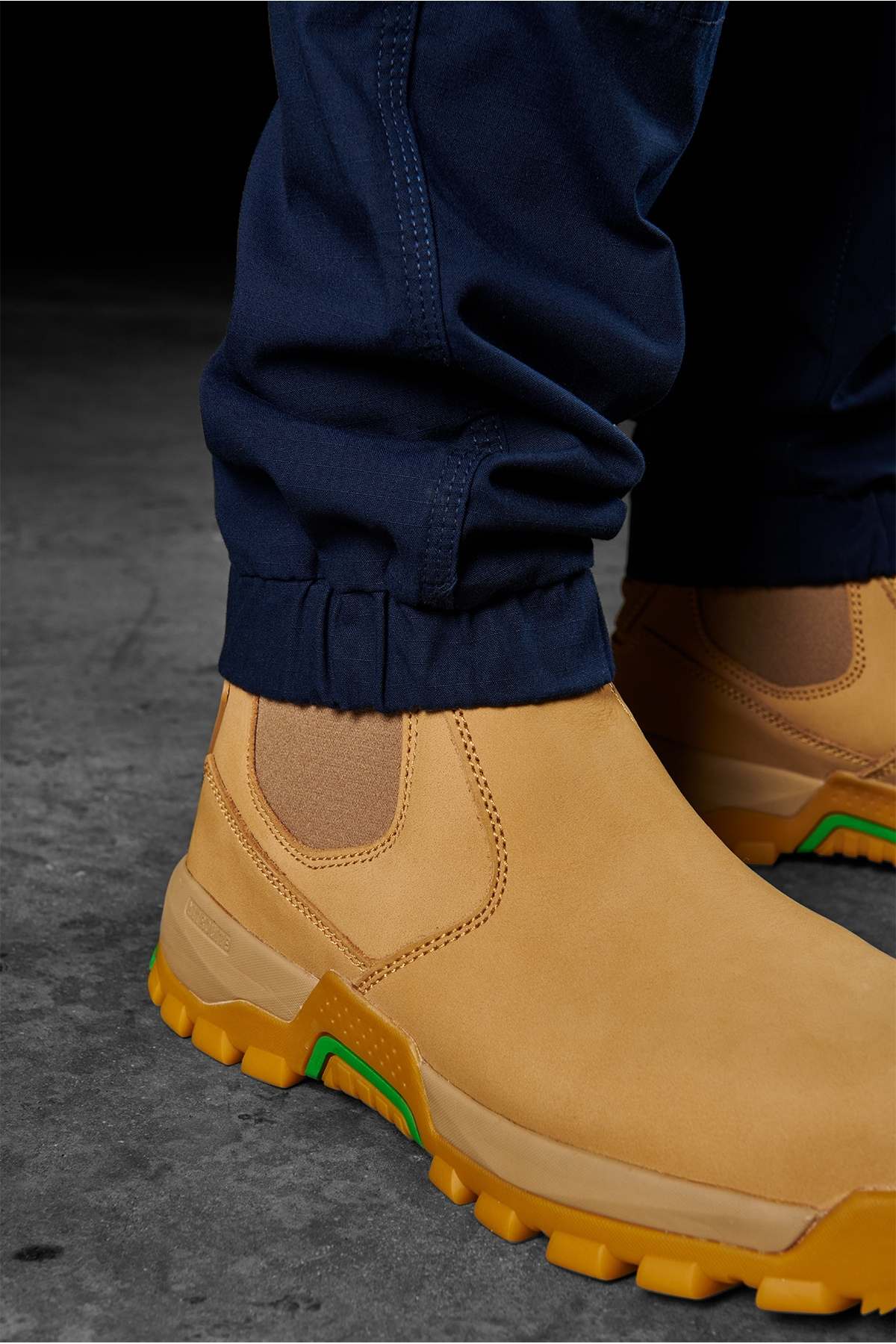 A close-up view of a person wearing yellow leather slip-on work boots with green accents and navy blue FXD WP-11 work pants. The photo highlights the rugged sole and elastic side panels of the boots, while cargo pockets on the FXD WP-11 pants add functionality. The background is a grey, textured surface.