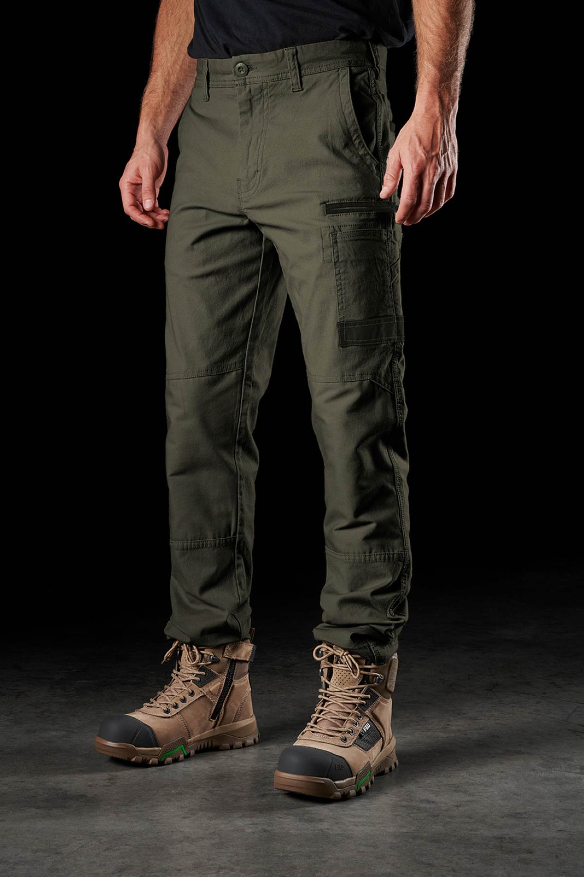 A person wearing FXD WP-3 olive green work pants with 360° stretch and brown lace-up boots stands on a dark surface against a black background, showing only the lower half of their body.