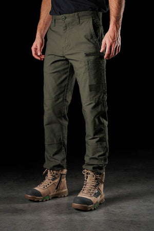 A person wearing FXD WP-3 olive green work pants with 360° stretch and brown lace-up boots stands on a dark surface against a black background, showing only the lower half of their body.