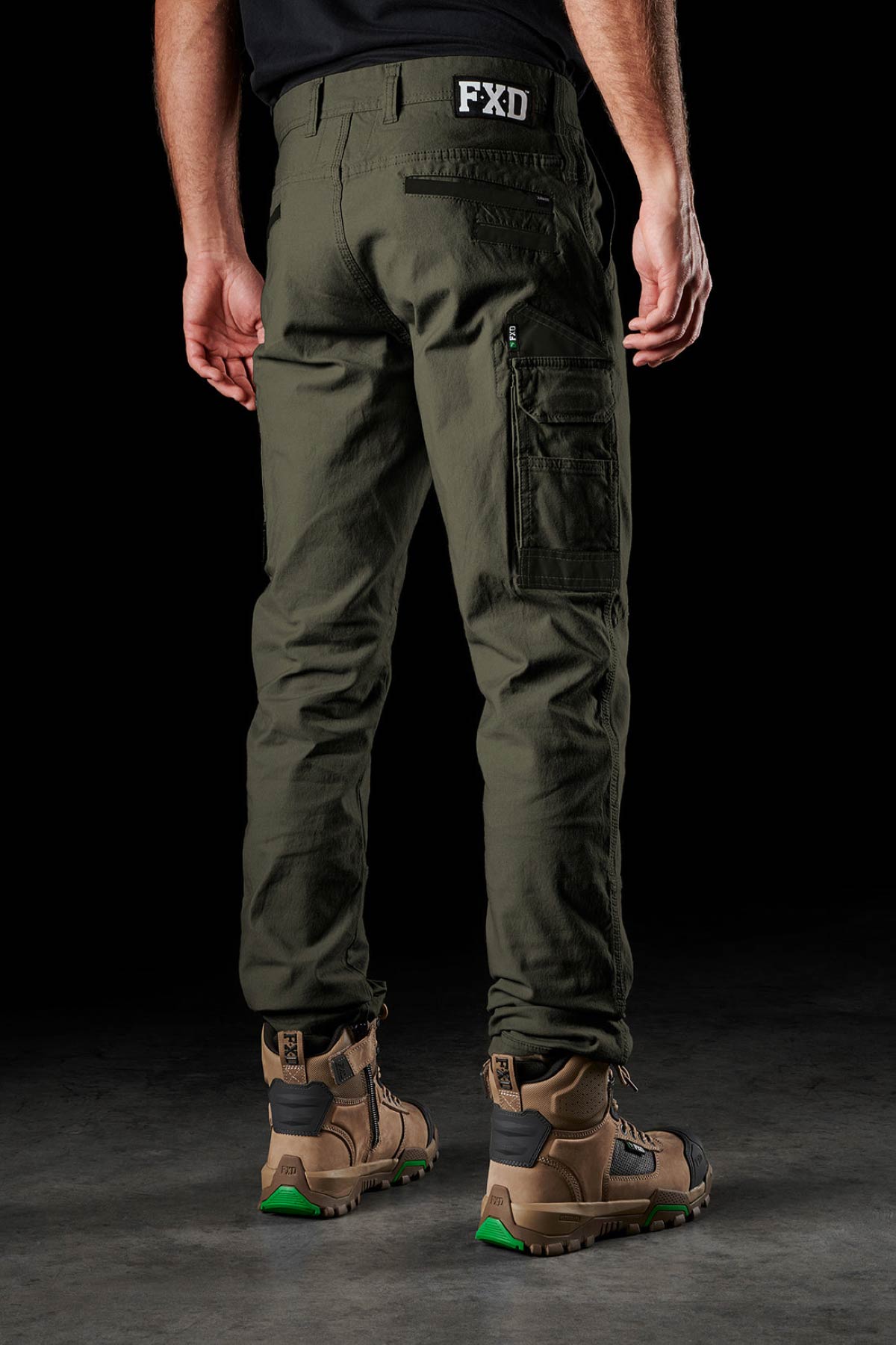 A person stands facing away in olive green FXD WP-3 360° stretch work pants and brown boots, highlighting the flexible back view of the pants against a dark background.