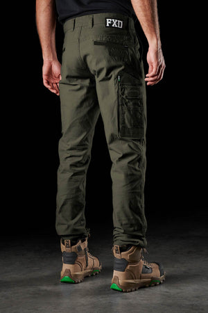 A person stands facing away in olive green FXD WP-3 360° stretch work pants and brown boots, highlighting the flexible back view of the pants against a dark background.