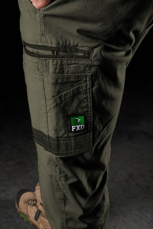 A close-up of a person wearing FXD WP-3 olive green work pants with 360-degree stretch and an FXD logo patch on the pocket, paired with tan work boots against a dark, blurred background.