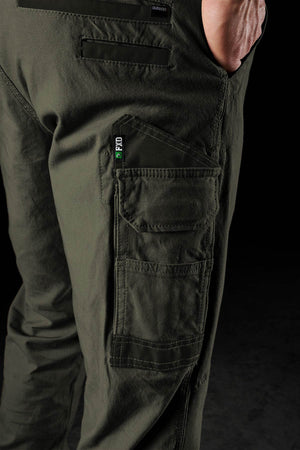 Close-up of a person wearing FXD WP-3 dark green work pants featuring multiple pockets, visible stitching, and a black and green “PRO” tag. The 360° stretch fabric ensures comfort as the hand rests in a pocket against a dark background.