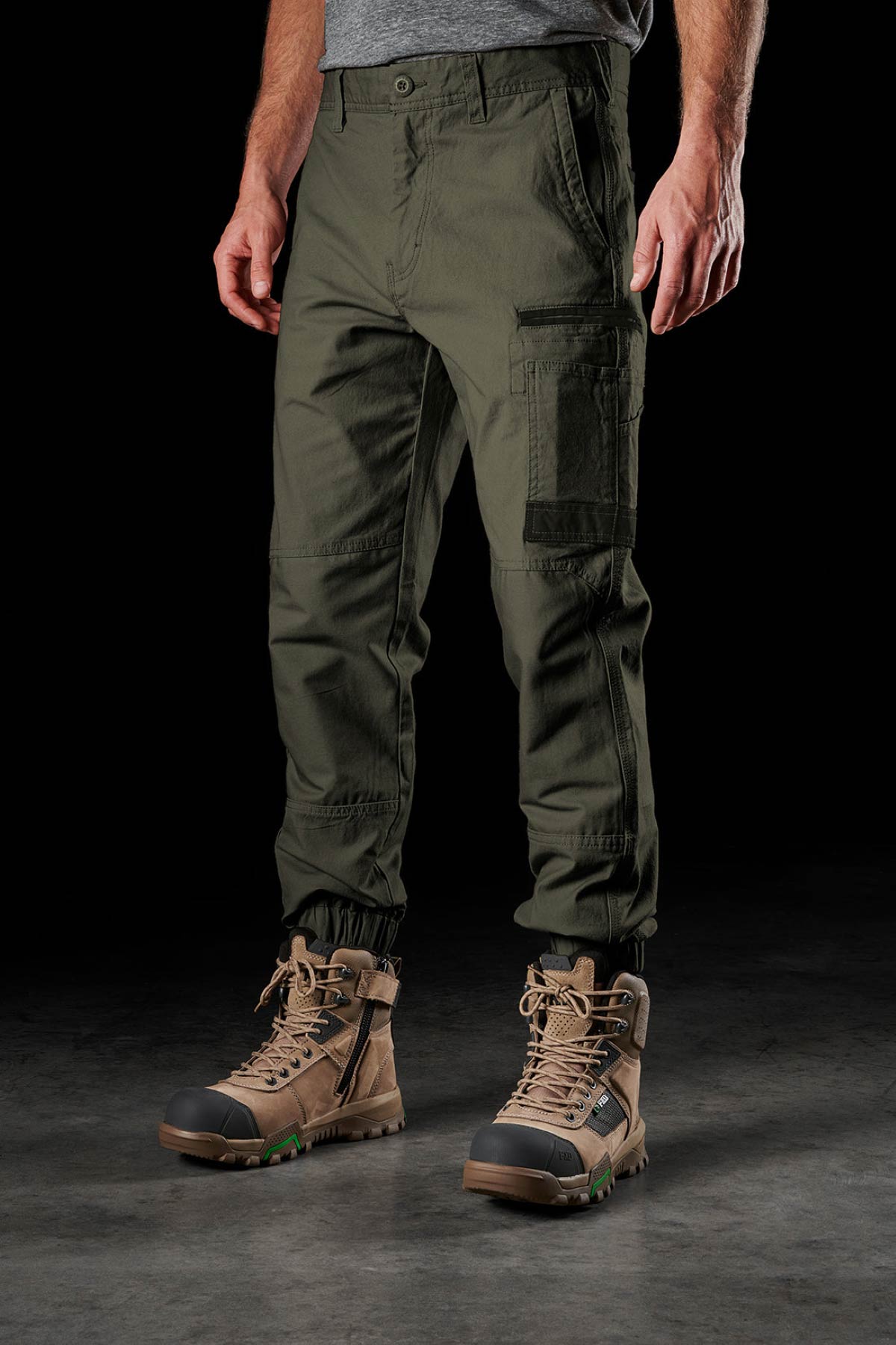 Someone in olive green FXD WP-4 Stretch Cuffed Work Pants and tan work boots stands against a dark background, with most of their upper body out of frame.