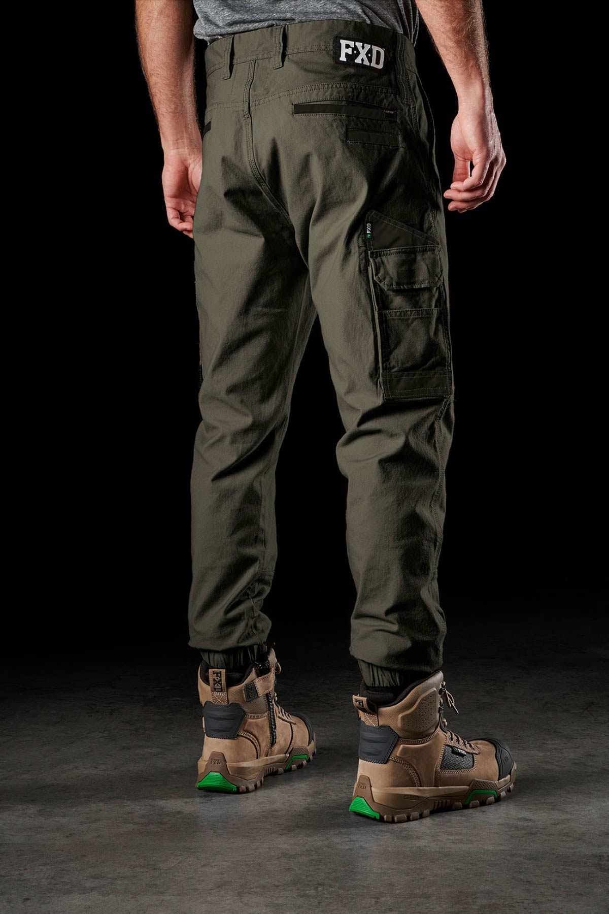 A person stands on a dark surface wearing olive green FXD WP-4 Stretch Cuffed Work Pants with multiple pockets and tan work boots. The "FXD" logo is visible near the waistband, while the upper body is mostly out of frame.